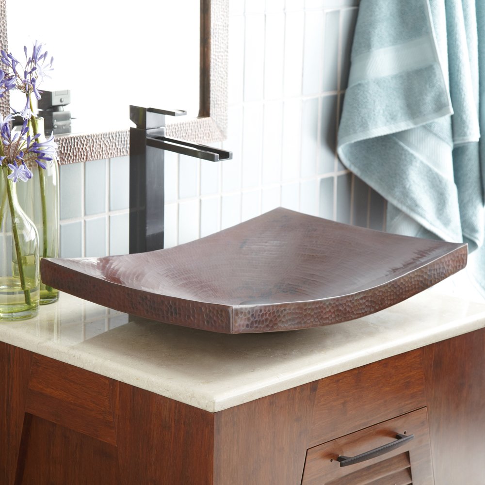 Antique copper vessel sink with curved rectangular shape and hammered texture, paired with tall black faucet on bathroom vani