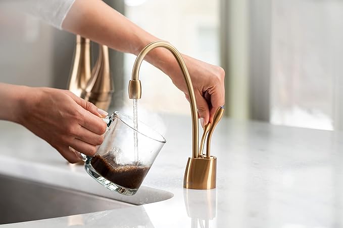 Brushed bronze instant hot and cold faucet dispensing dark liquid into a glass held over a sink.