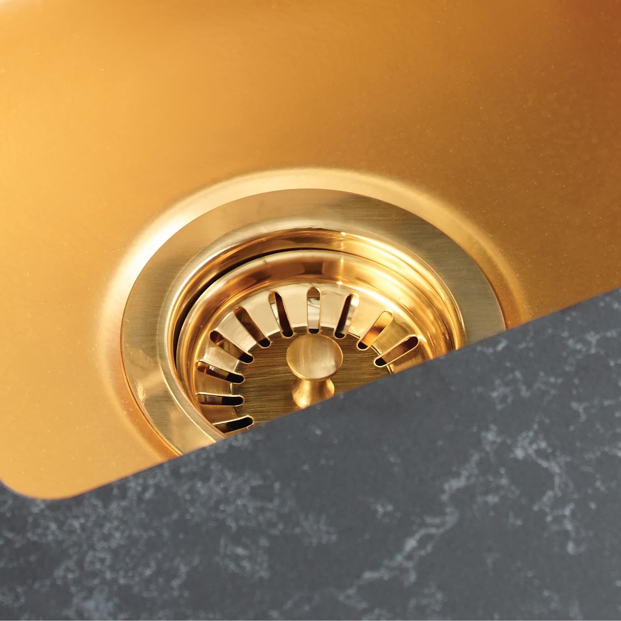 Matte black basket strainer with gold-tone metal interior and perforated drainage slots, installed in dark countertop.