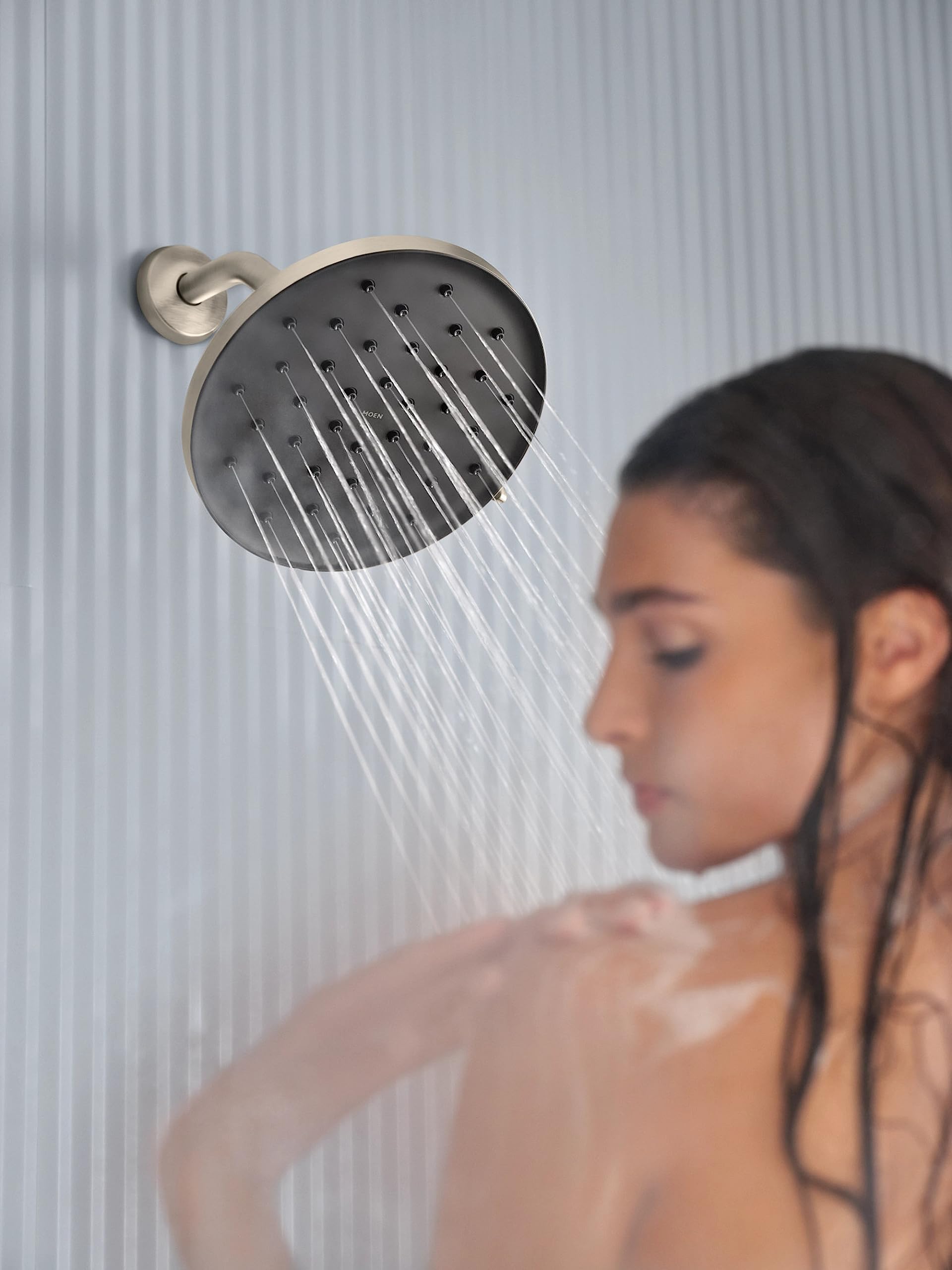 Brushed nickel round rainshower head with multiple spray nozzles mounted on wall with curved arm, dispensing water.
