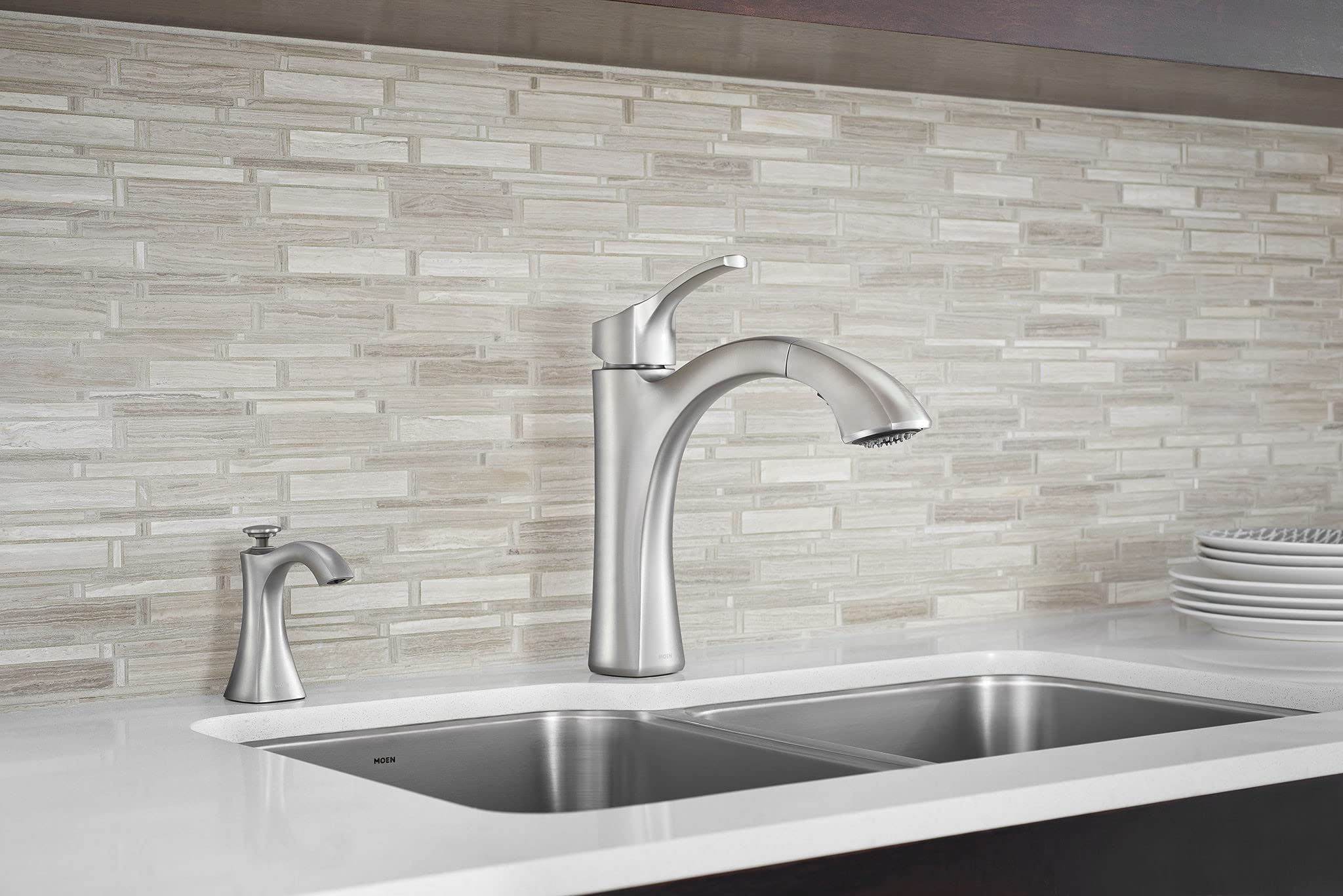 Chrome soap dispenser mounted on white kitchen countertop next to stainless steel faucet and double sink.