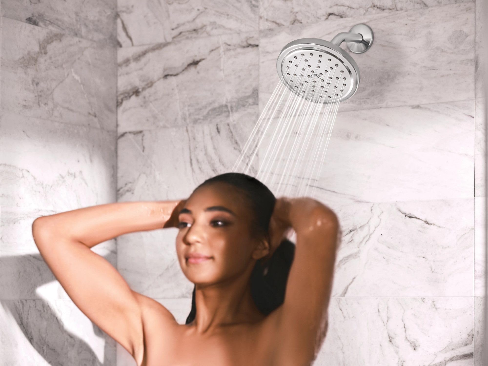 Chrome showerhead with water spray mounted on marble wall with person showering beneath it.