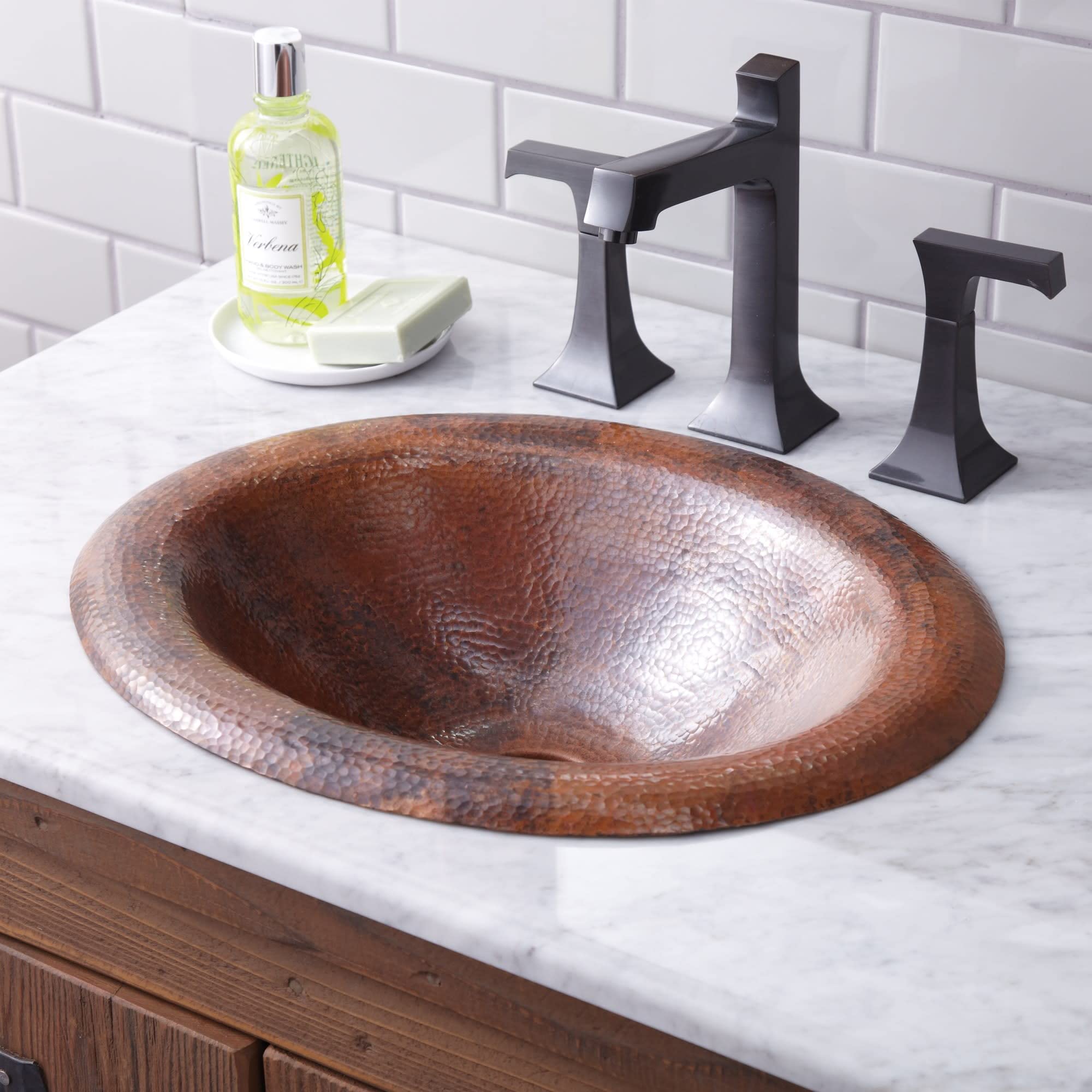 Oval copper sink with hammered finish on marble countertop, paired with black two-handle faucet and subway tile backsplash.