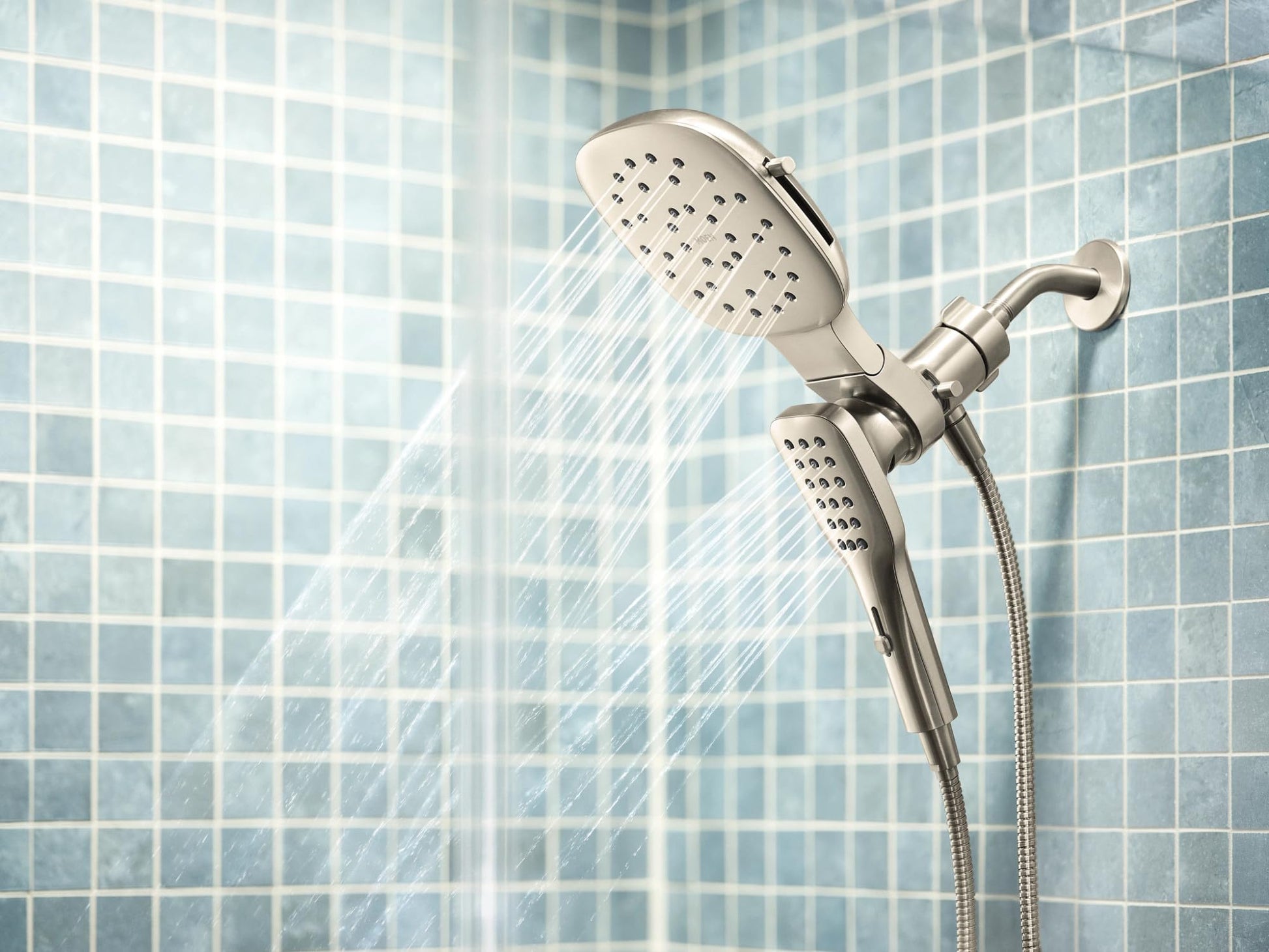 Brushed nickel square rain shower head with detachable hand shower and hose mounted on tile wall with water spray visible.