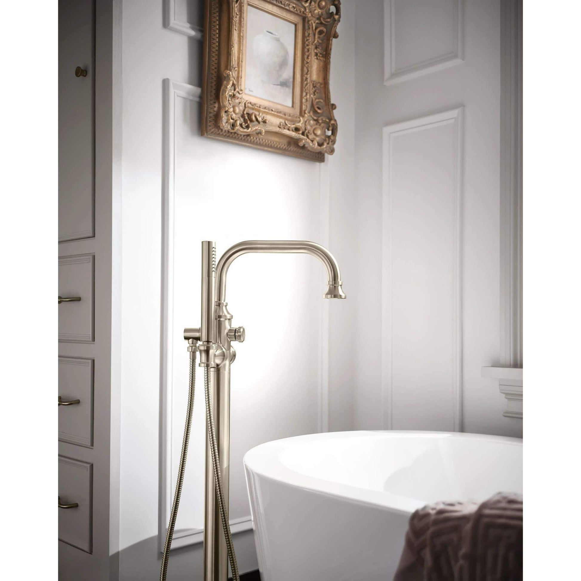 Matte black freestanding tub filler with curved spout, single handle, and handheld shower on hose next to white soaking tub.