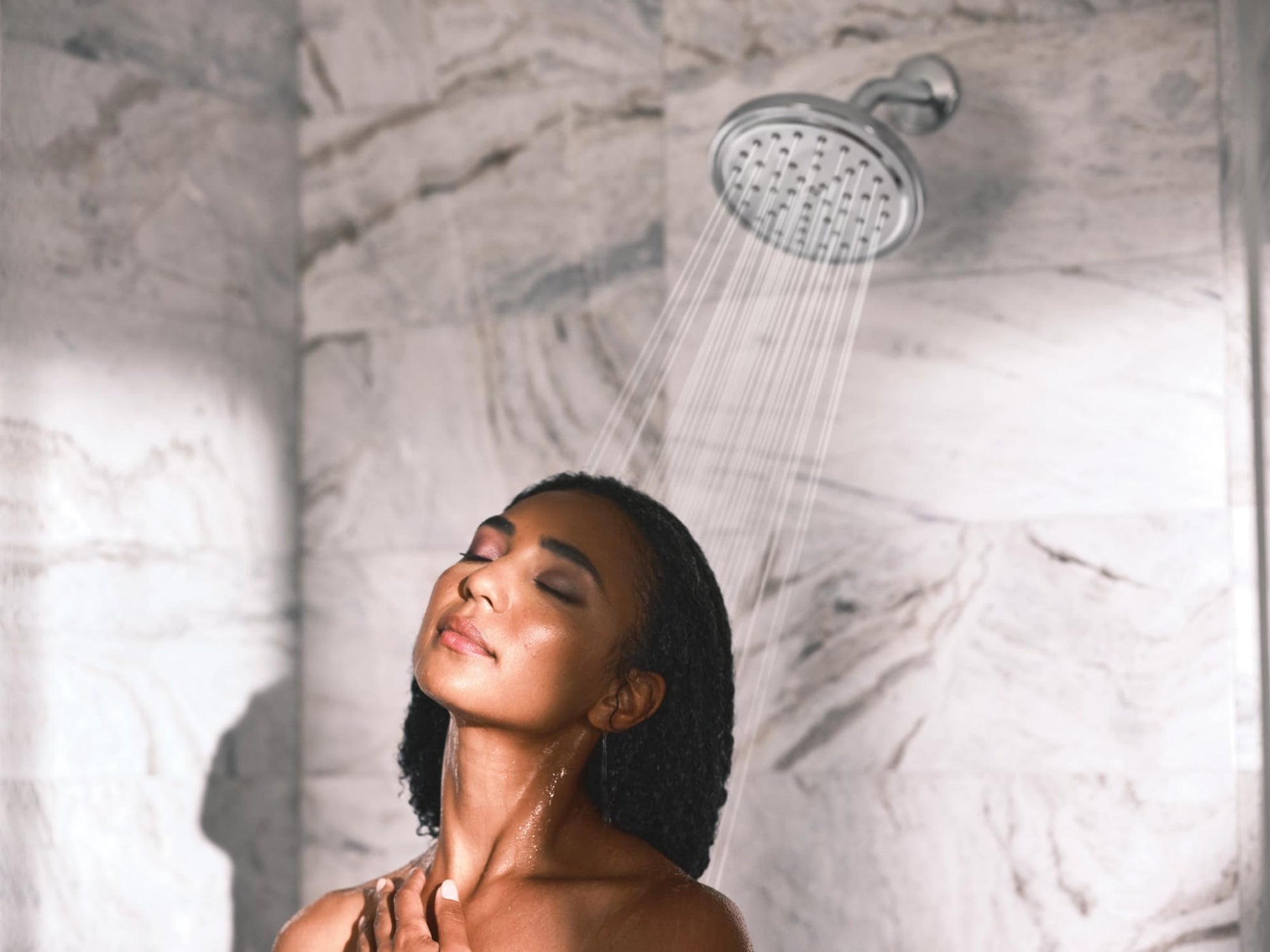 Chrome showerhead with water streams spraying down on a person's face in a marble-tiled shower.