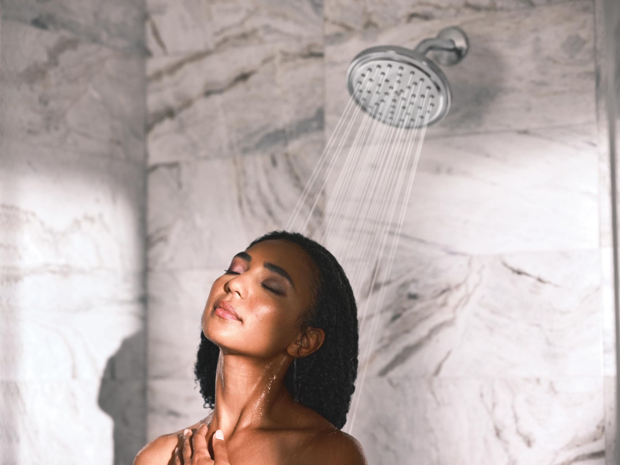 Chrome showerhead with water streams spraying down on a person's face in a marble-tiled shower.