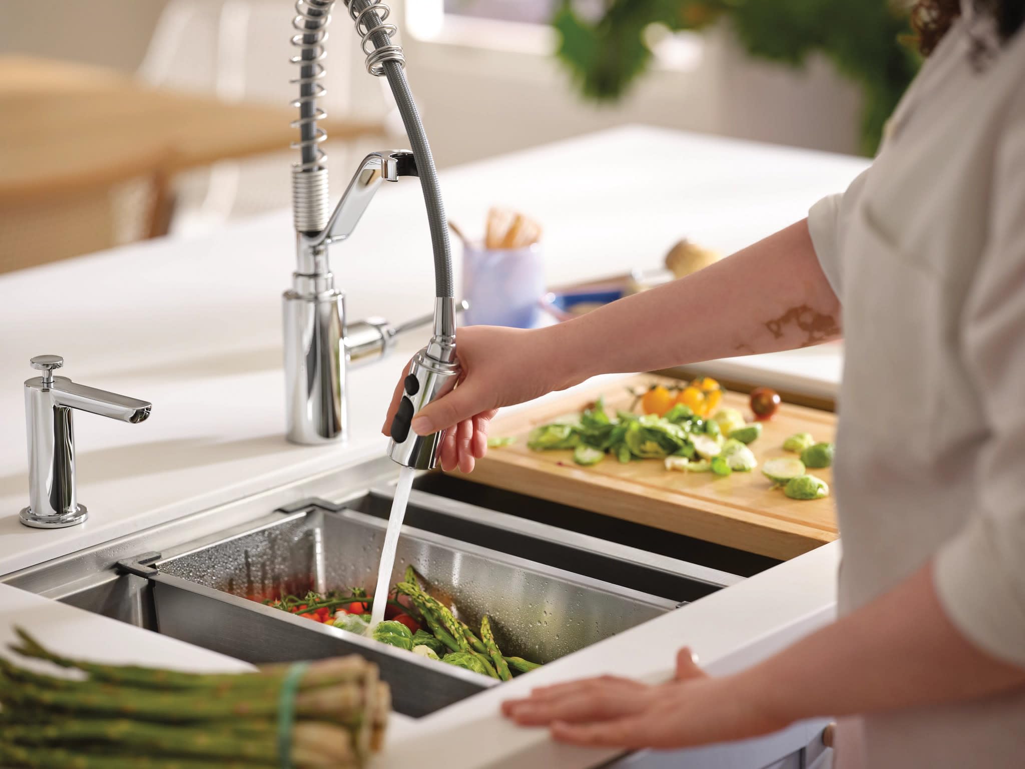 Stainless steel double bowl undermount kitchen sink with pull-down faucet, drying rack, and cutting board in use with vegetab