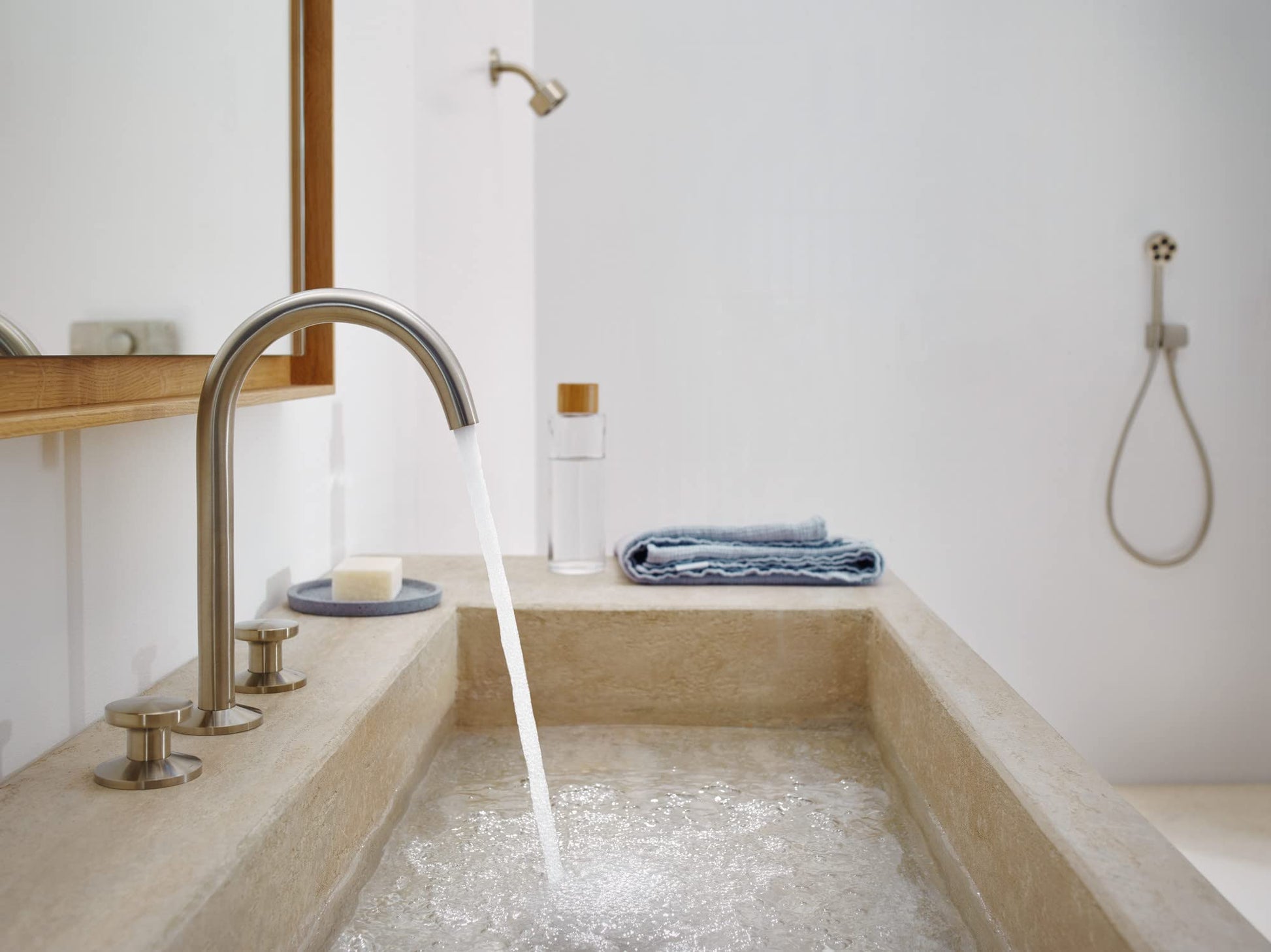 Brushed nickel shower head mounted on white bathroom wall above soaking tub with water running.