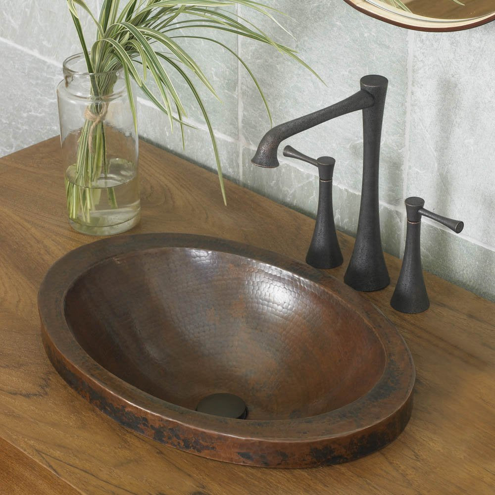 Hammered copper oval bathroom sink with dark patina finish installed on wood countertop with black faucet.
