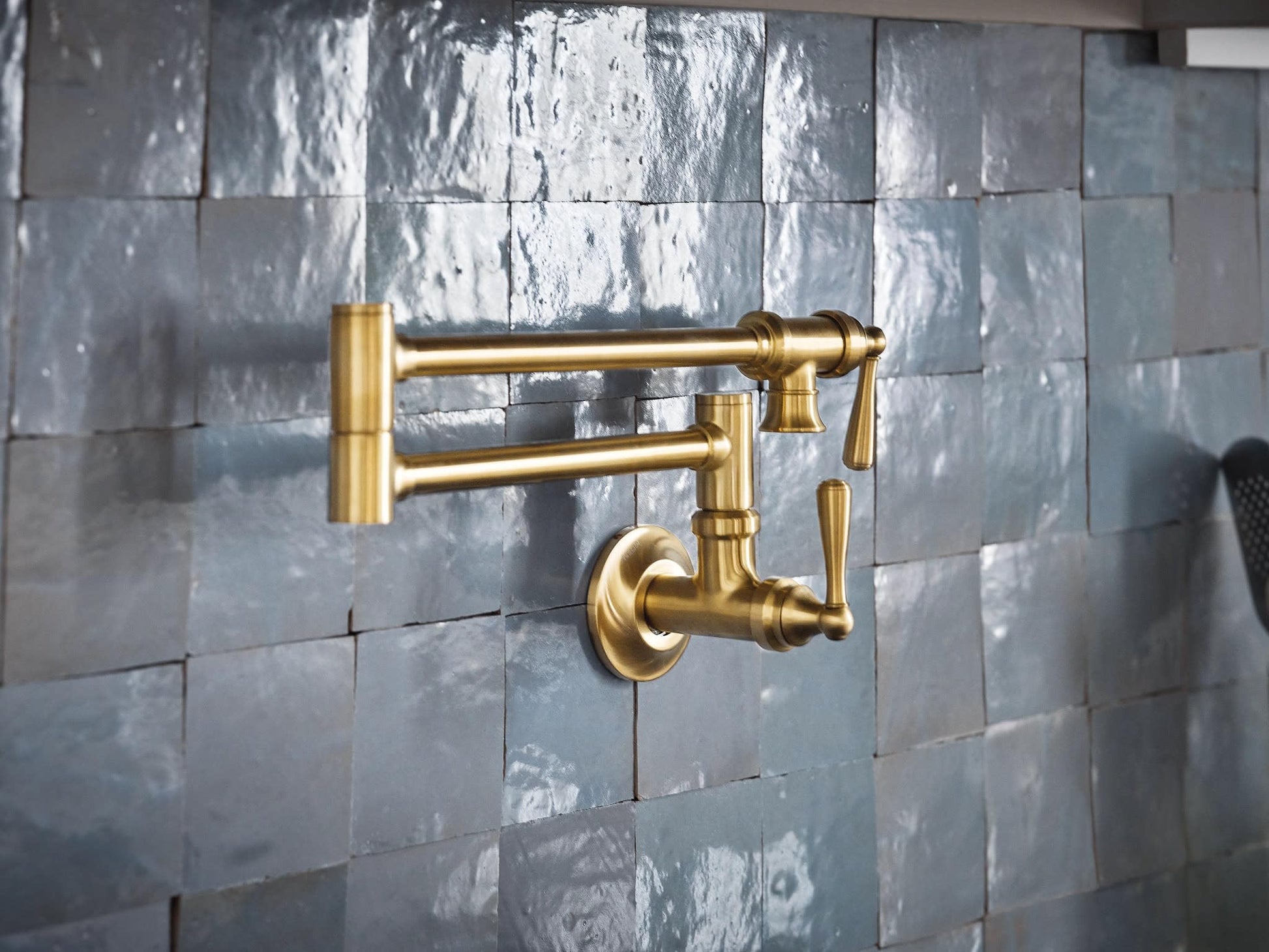 Brushed gold wall-mounted pot filler faucet with articulating arm and lever handle on tile backsplash.
