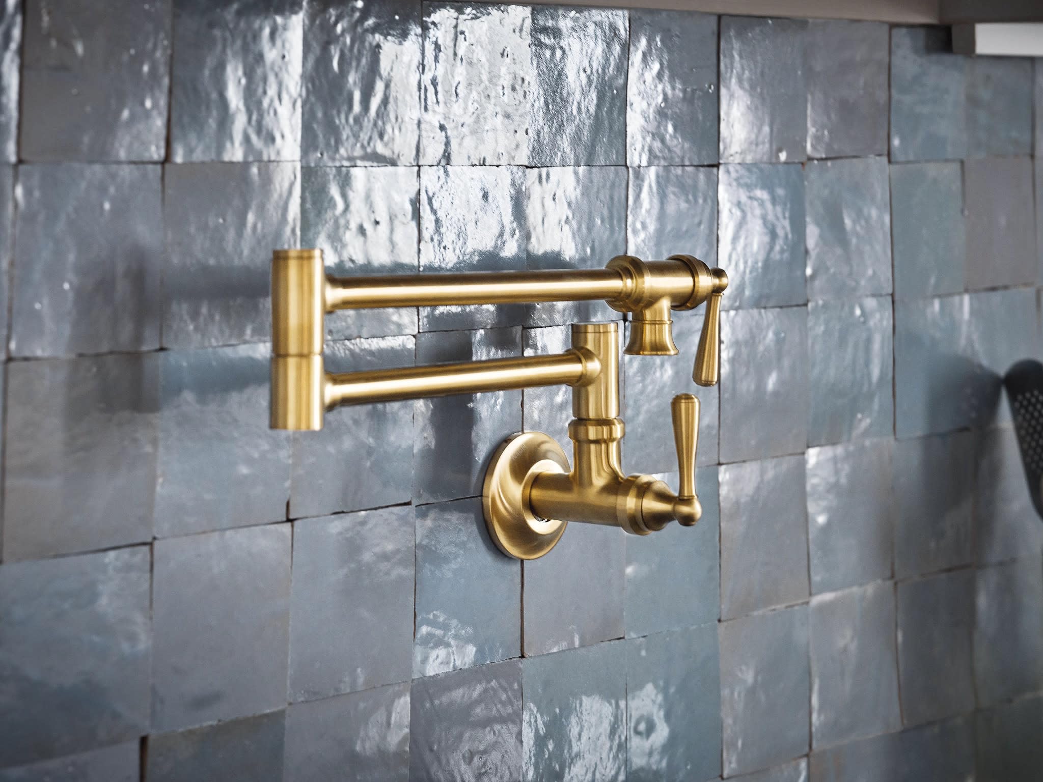 Brushed gold wall-mounted pot filler faucet with articulating arm and lever handle on tile backsplash.