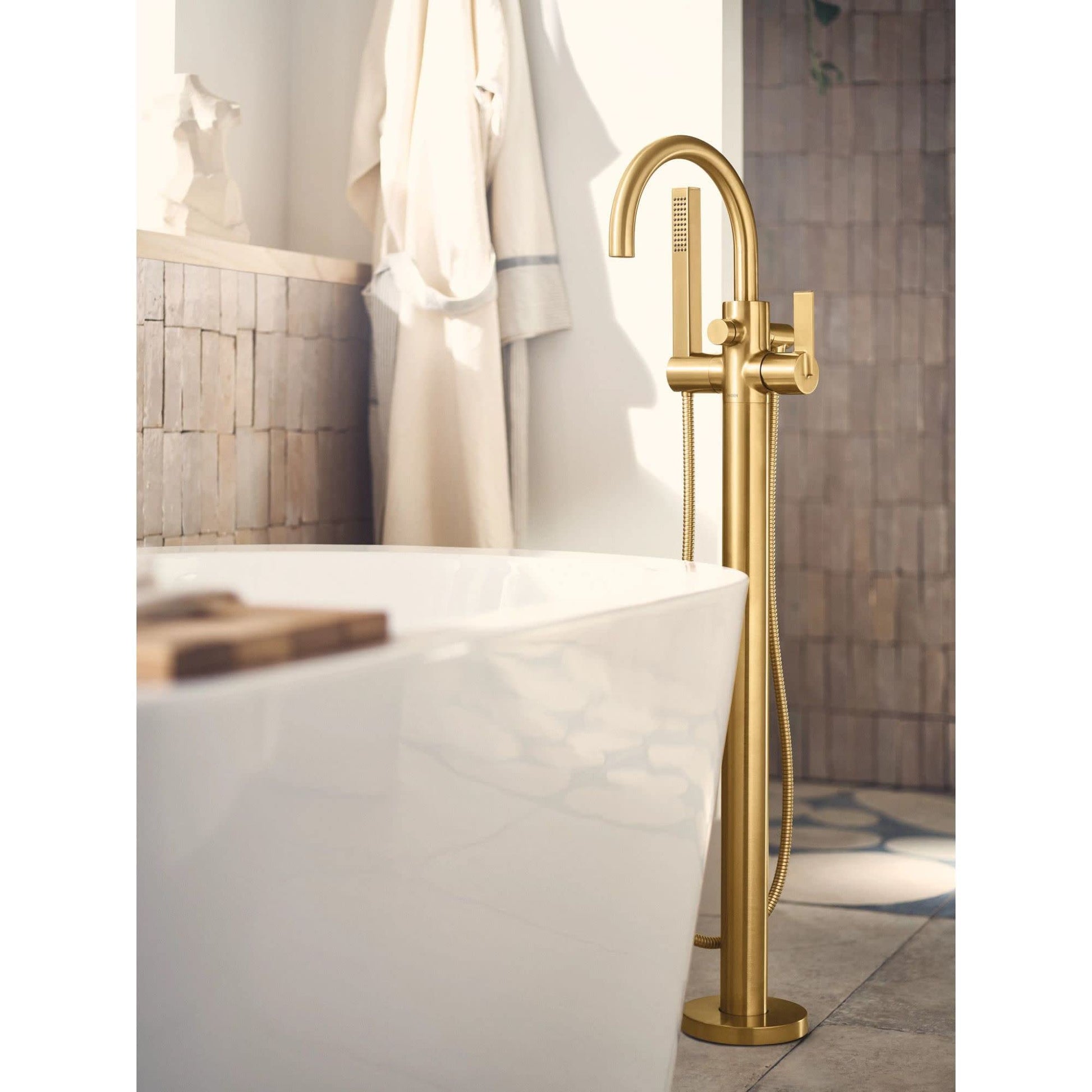 Brass floor-mount tub filler with curved spout, single handle, and attached hand shower hose next to white freestanding batht