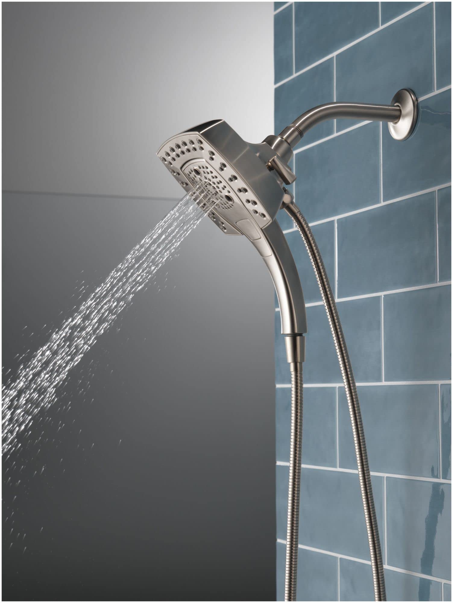 Chrome shower head with curved arm mounted on tile wall, water spraying from multi-hole perforated face with handheld wand at