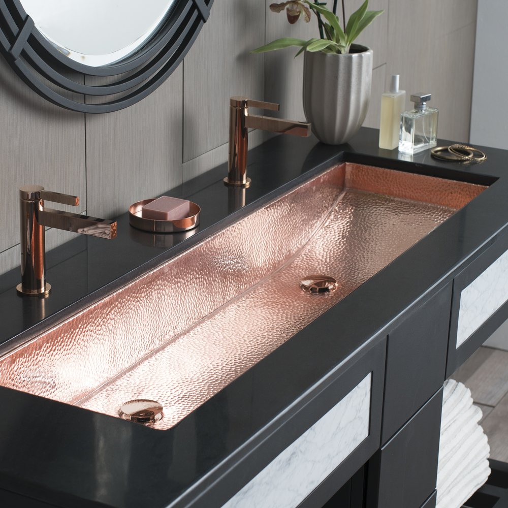 Polished copper trough sink with hammered surface texture and two drain holes mounted in black countertop.