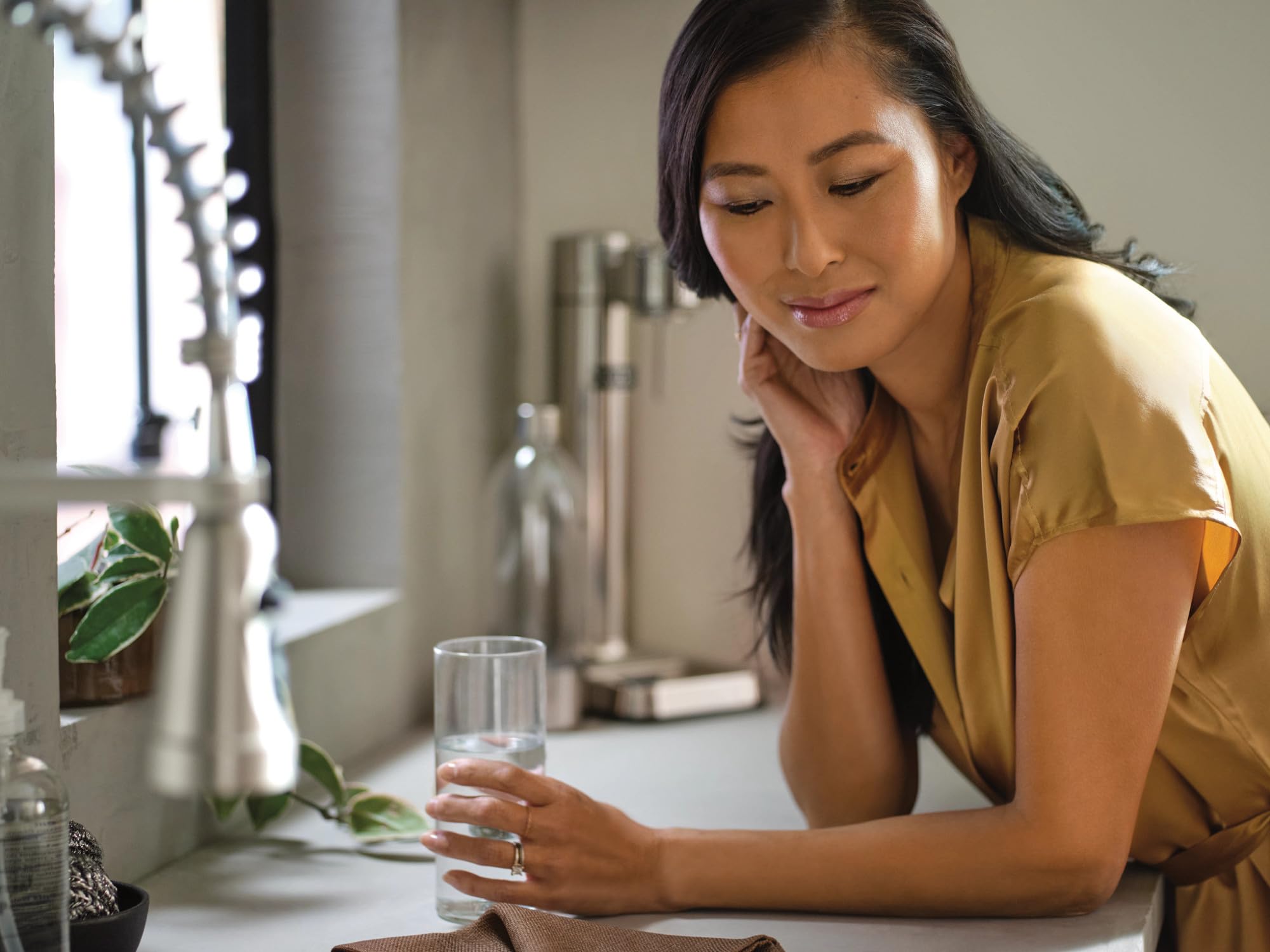 Woman in yellow shirt holding glass of water at kitchen sink with filtration system installed underneath.