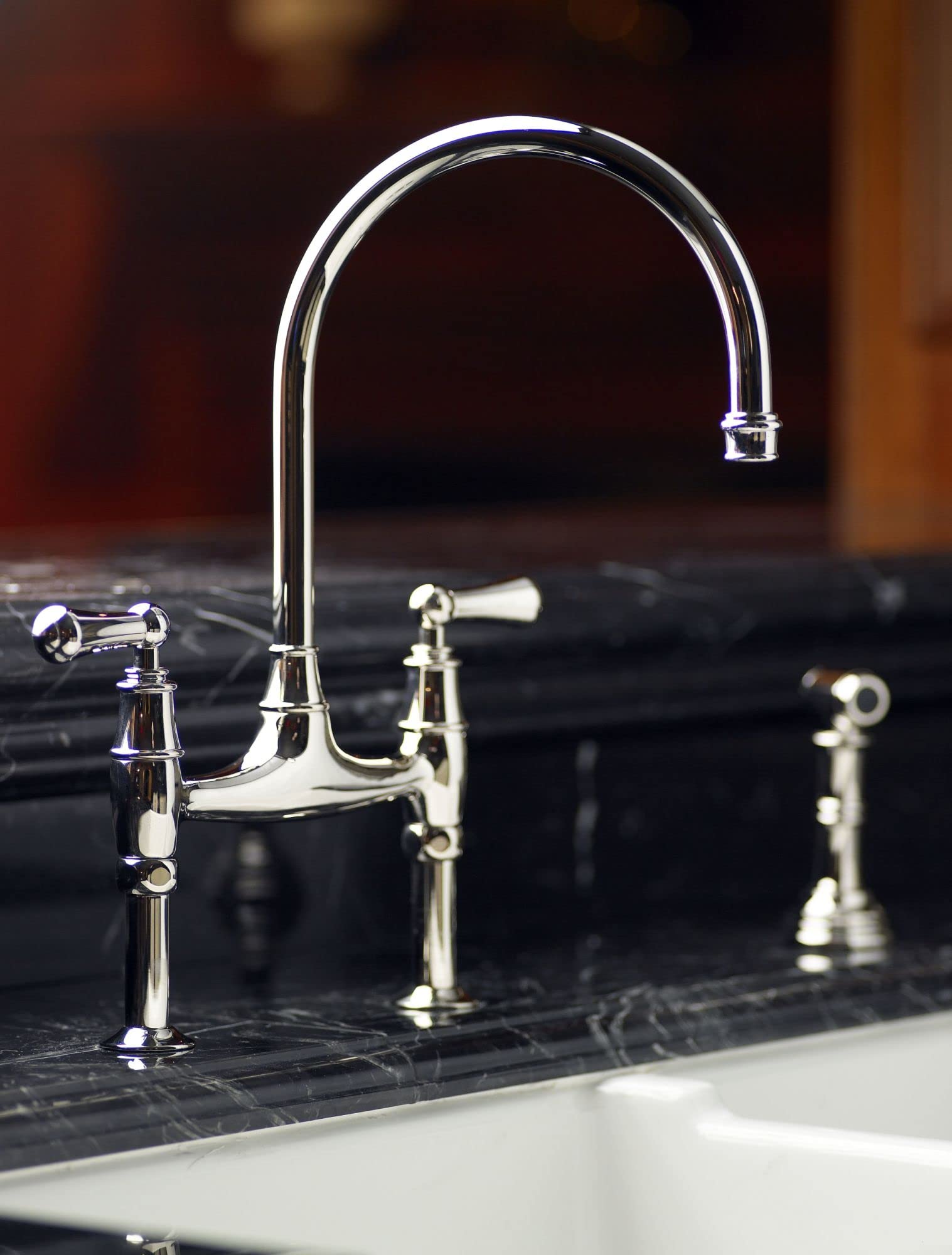 Satin English gold bridge kitchen faucet with curved spout and side spray on dark marble countertop.