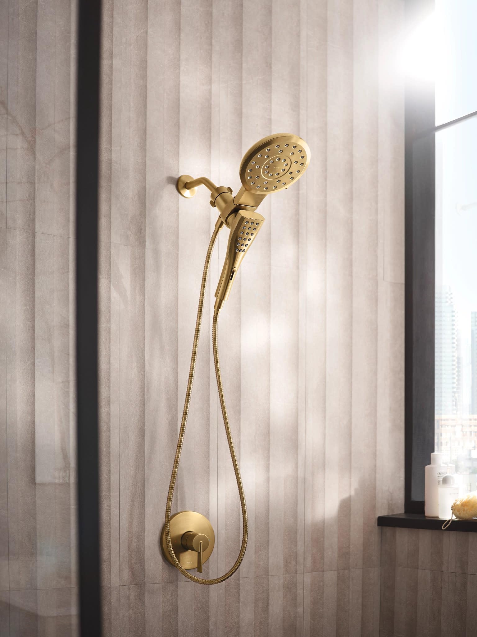Brushed gold dual shower head combo with handheld and wall-mounted spray heads connected by flexible hoses on tile wall.