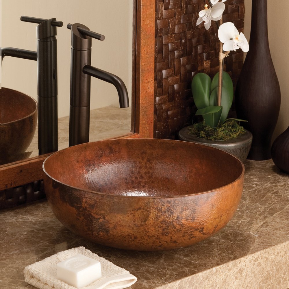 Round hammered copper vessel sink with textured surface, displayed with black faucets and bathroom accessories.