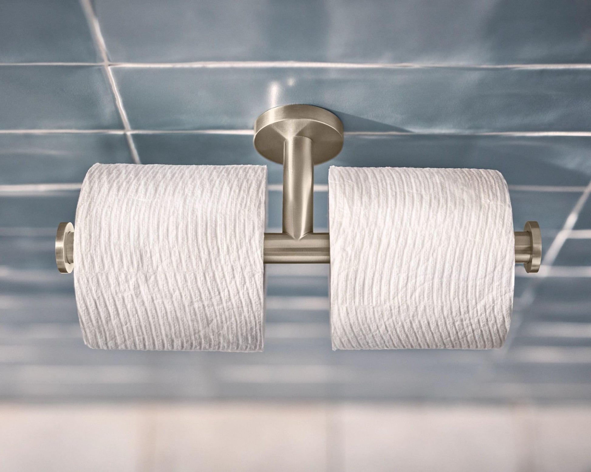 Brushed nickel double toilet paper holder with two white rolls mounted on tiled bathroom wall.