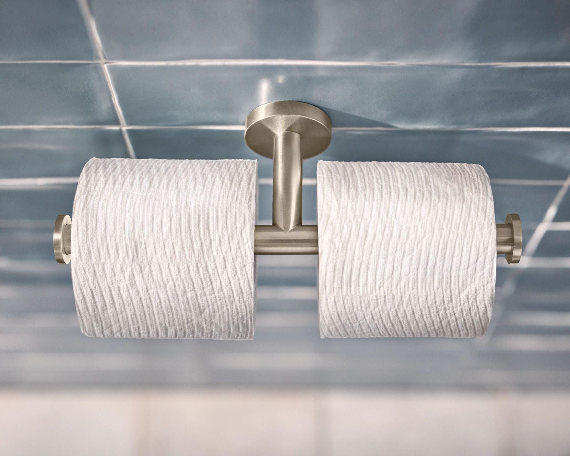 Brushed nickel double toilet paper holder with two white rolls mounted on tiled bathroom wall.