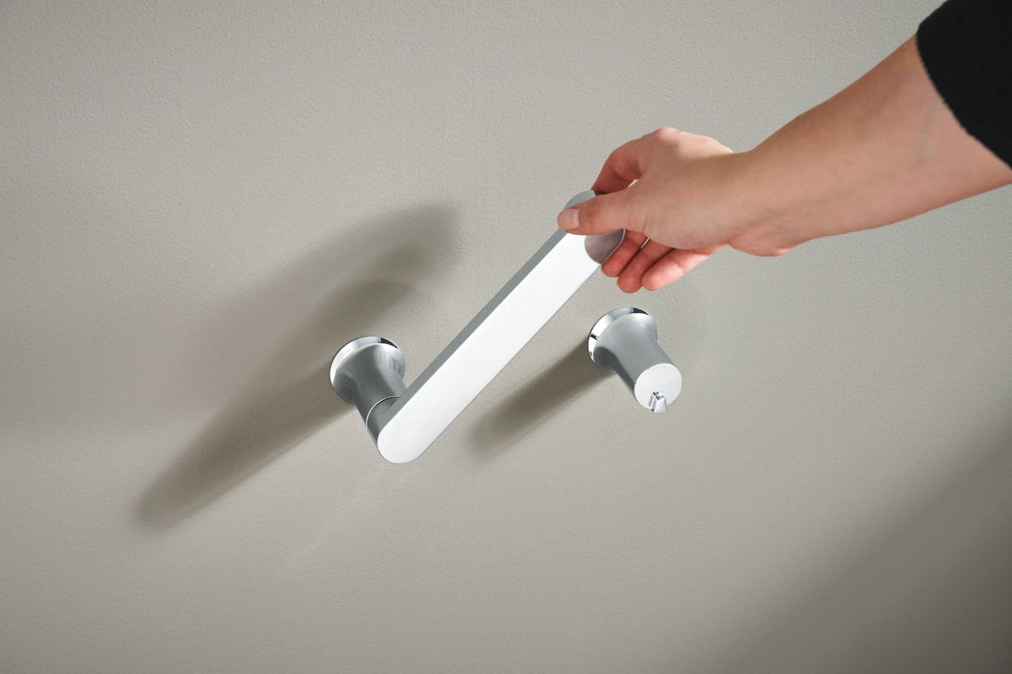 Chrome and white pivoting toilet paper holder mounted on wall with hand pulling down the rotating arm.