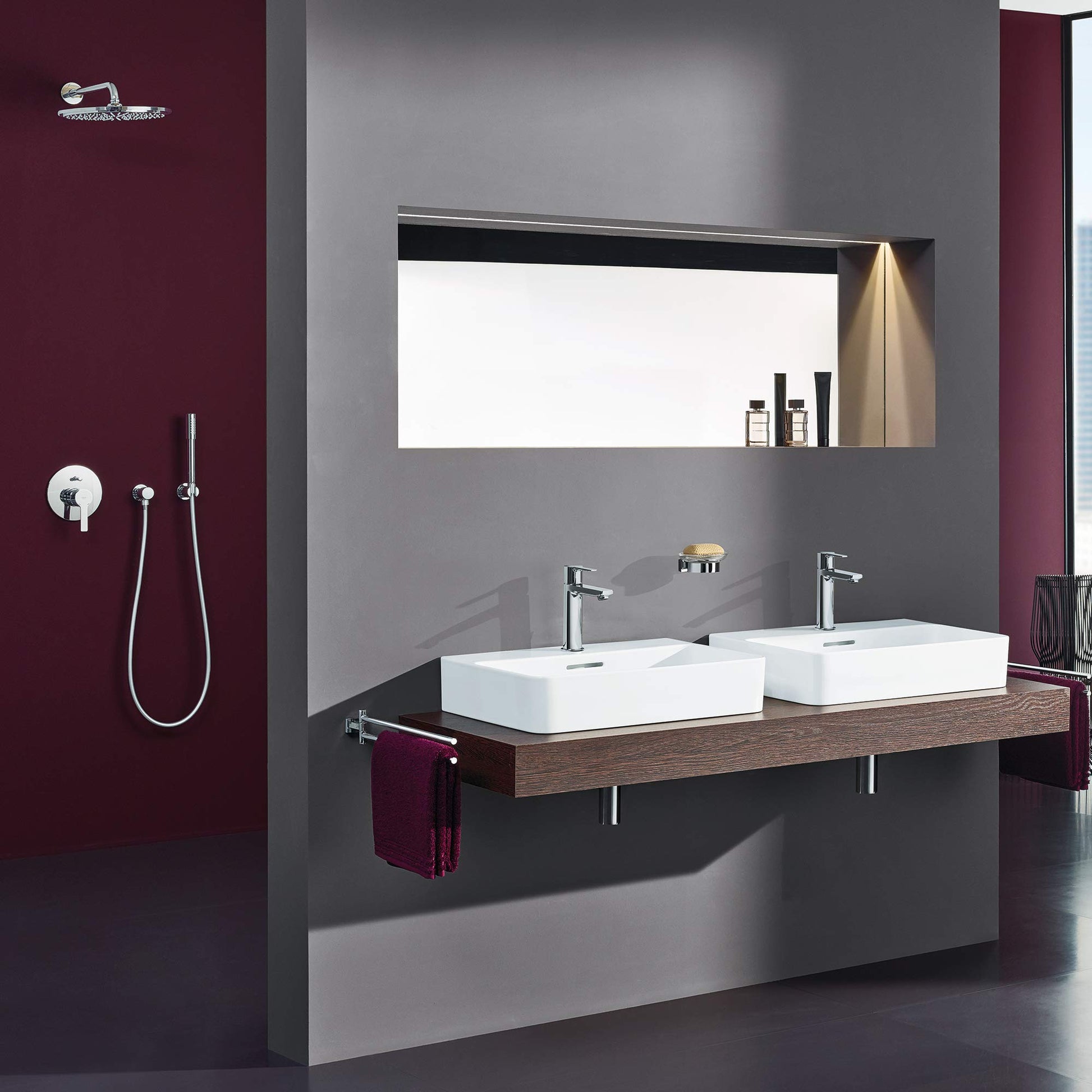 Chrome 3-way diverter trim with round handle and linear spout mounted on burgundy bathroom wall above double vessel sinks.