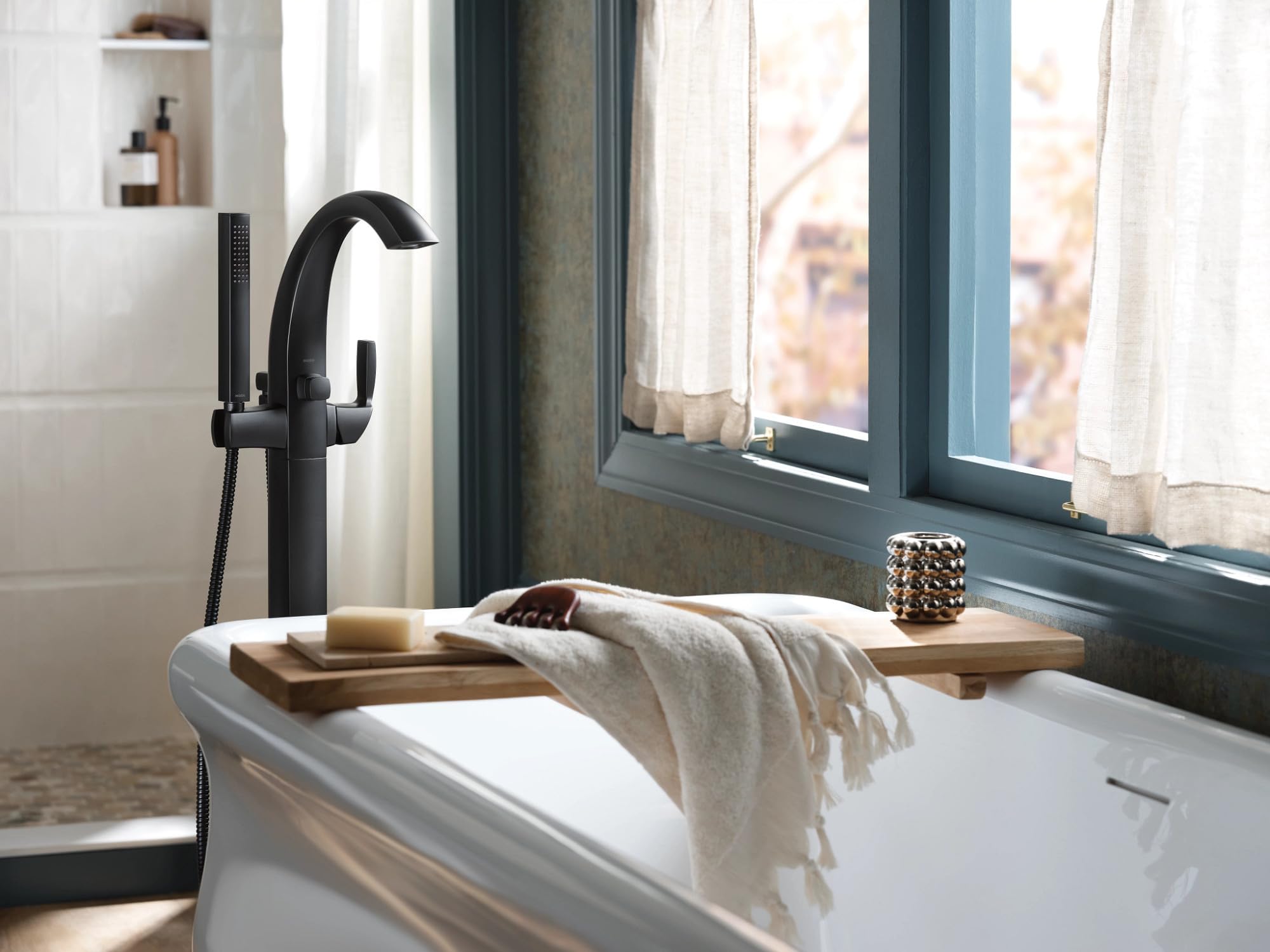Matte black freestanding tub filler with curved spout, single handle, and detachable handshower mounted on white bathtub.