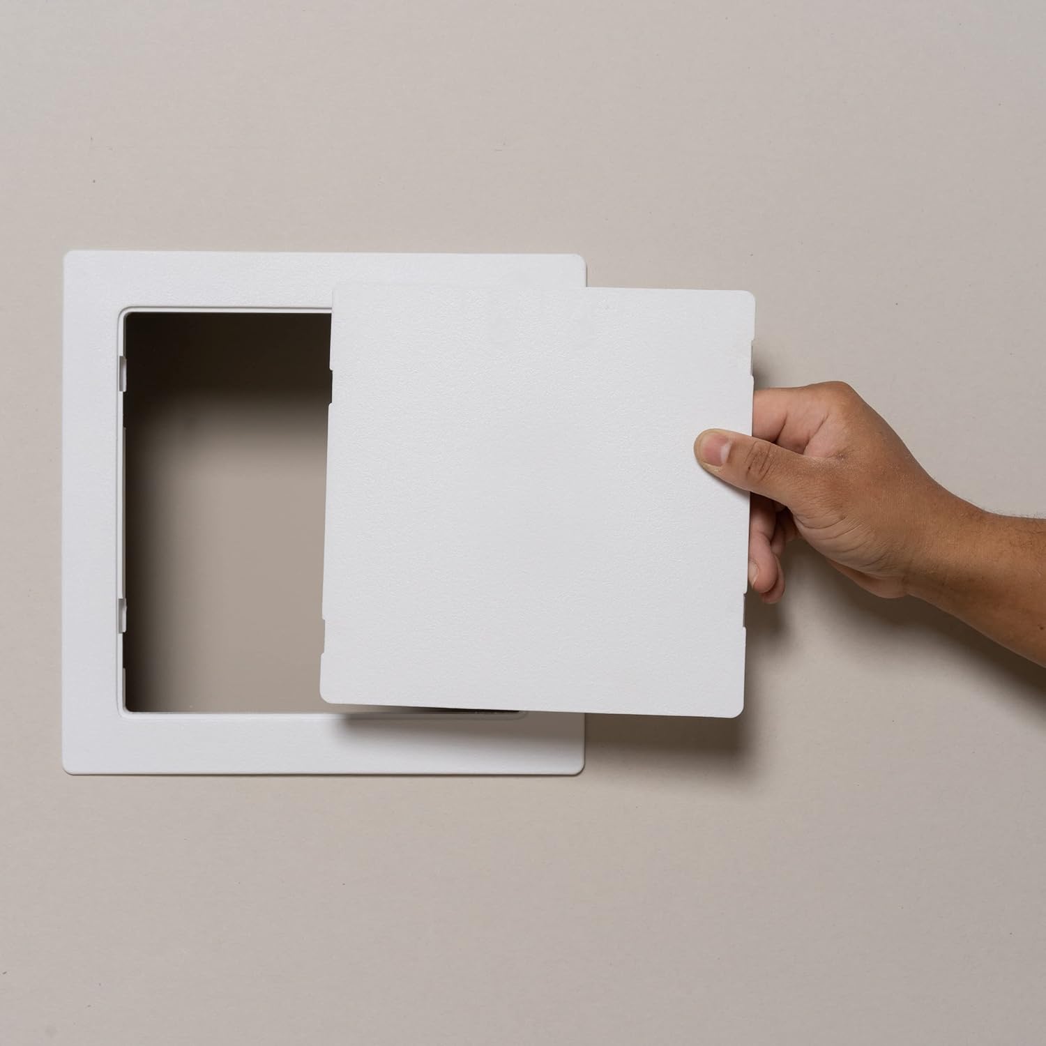 White plastic access panel with hinged door, shown open and held by a hand against a neutral background.