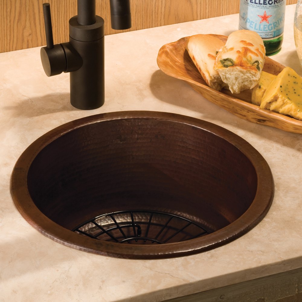 Hammered copper round bar sink with dark patina finish, black faucet, and drain basket installed in light countertop.