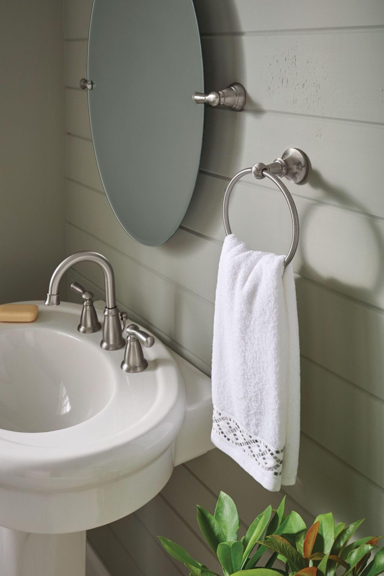 Brushed nickel wall-mounted towel ring holding a white towel next to a bathroom mirror and sink.