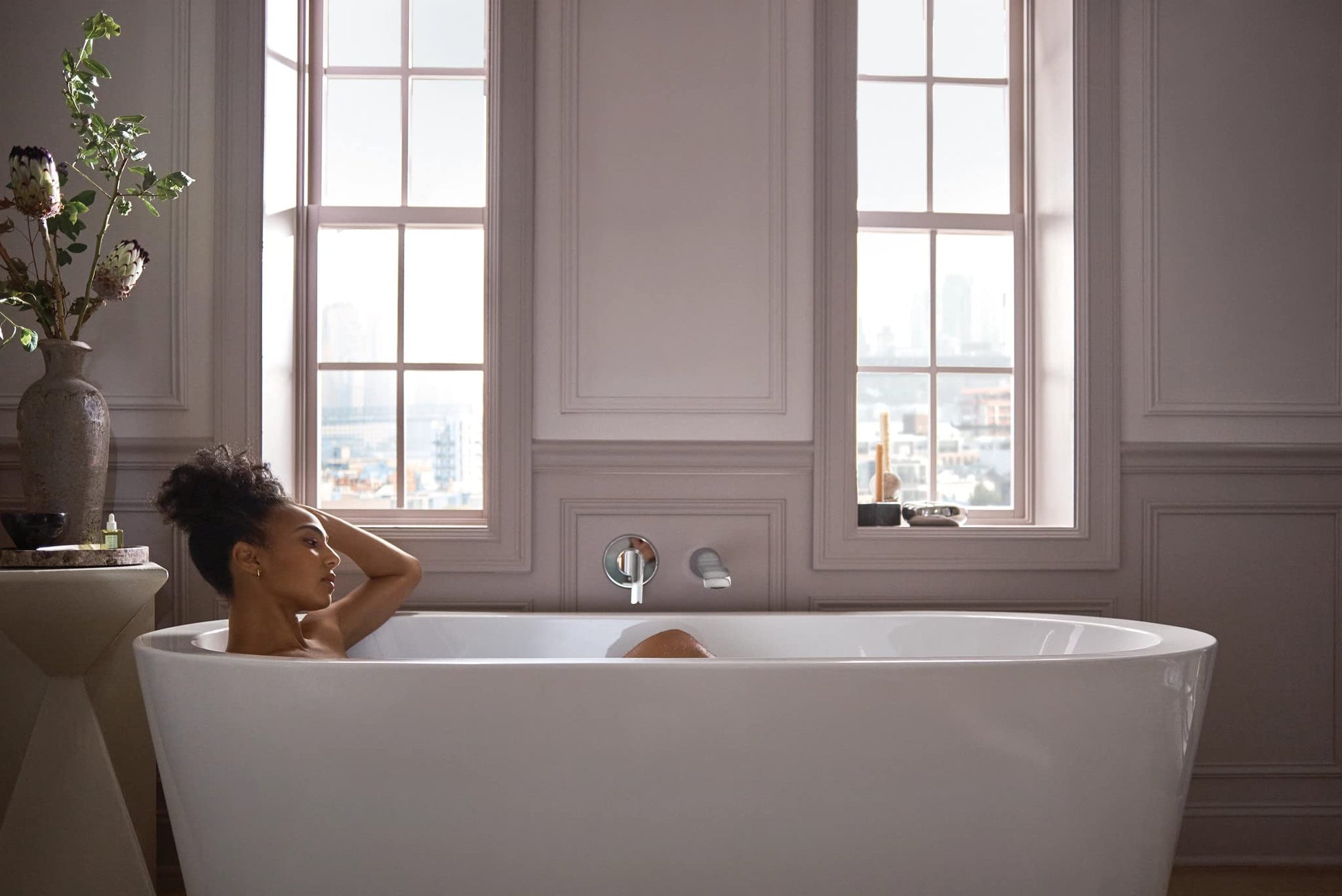 Chrome wall-mounted tub faucet with single handle and extended spout installed above white freestanding bathtub.