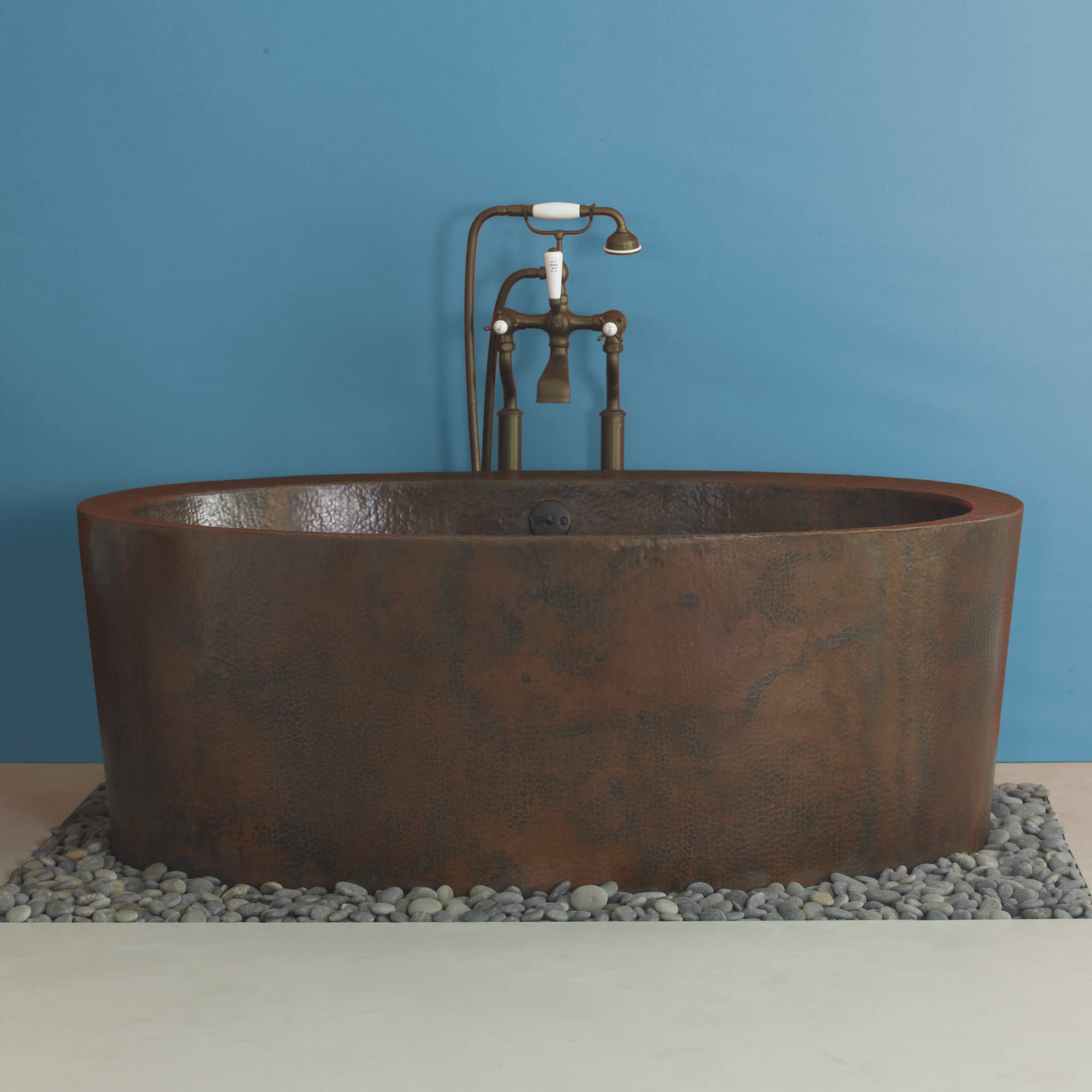 Freestanding oval hammered copper bathtub with vintage bronze faucet and hand shower, displayed on river stones.
