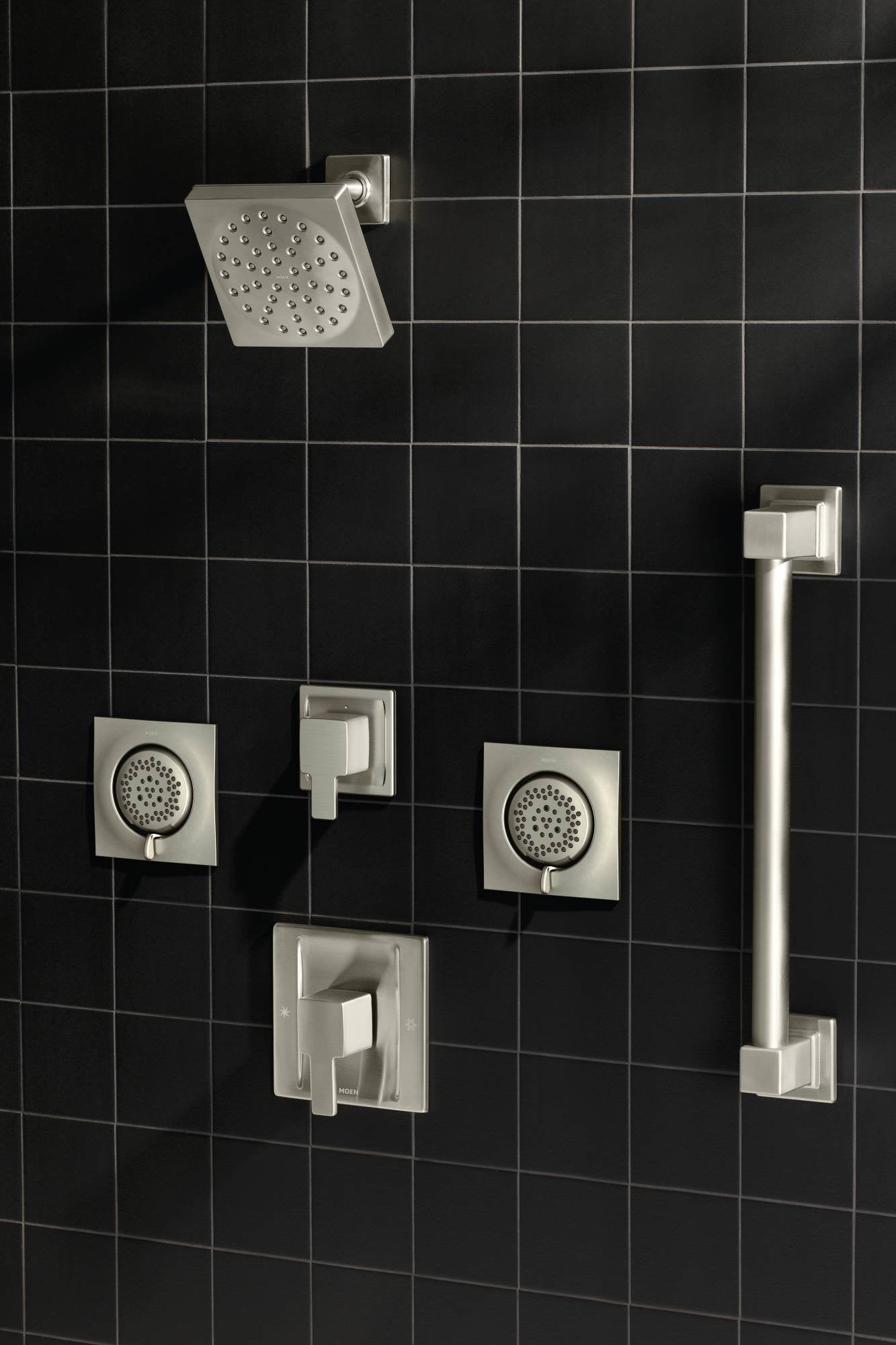 Brushed nickel shower system with square head spray, two body jets, valve trim, and grab bar mounted on black tile wall.