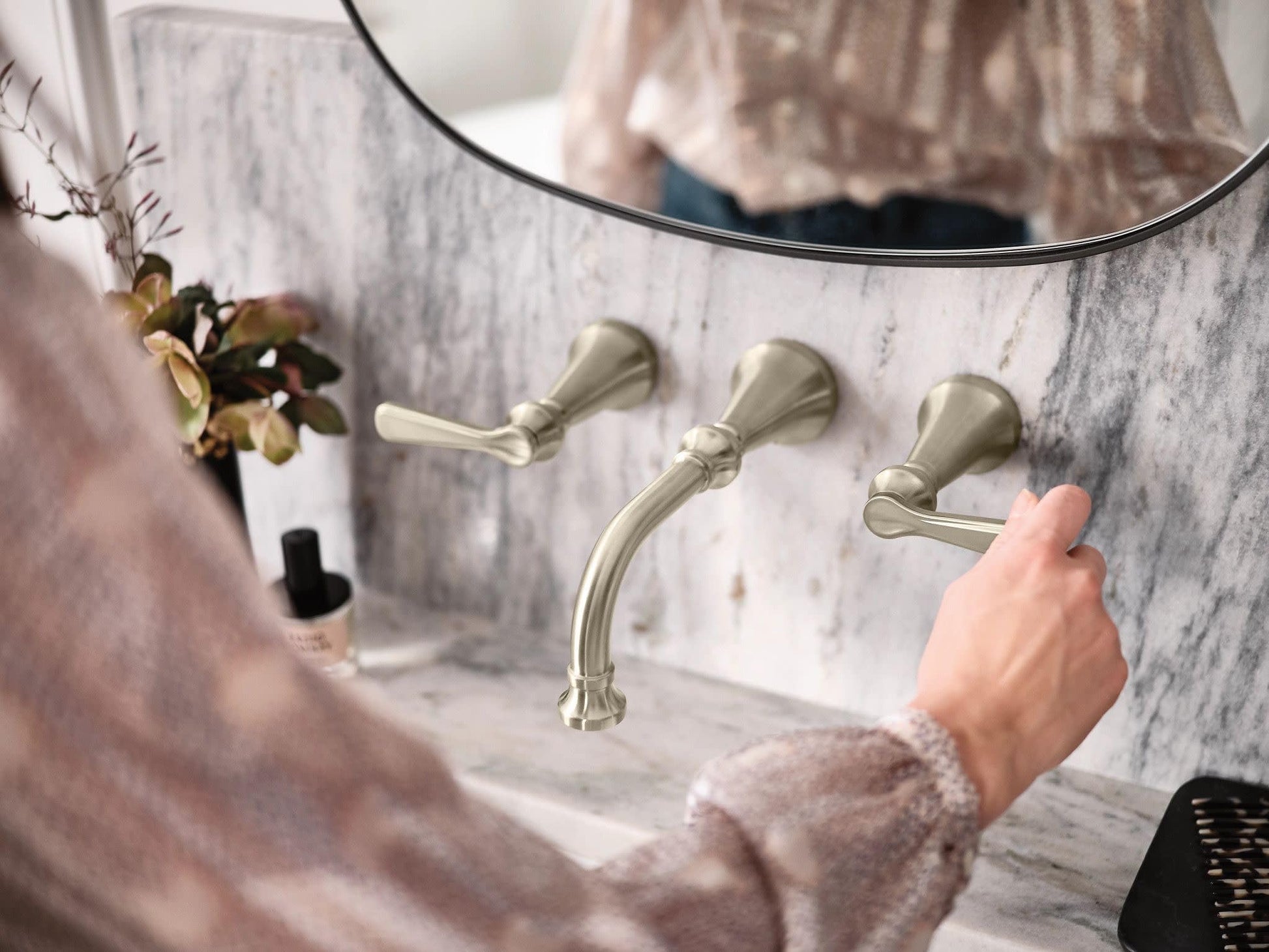 Brushed nickel wall-mount bathroom faucet trim with three lever handles and curved spout installed on marble wall.