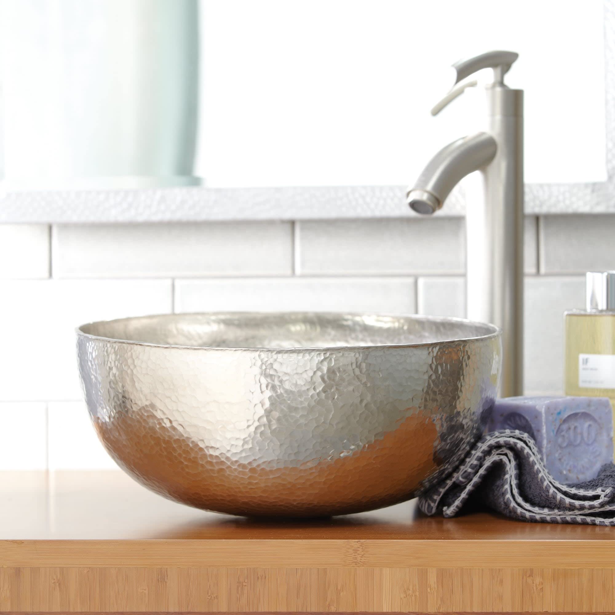 Hammered metal vessel sink in antique finish with curved tall faucet and spray, displayed on wooden counter.