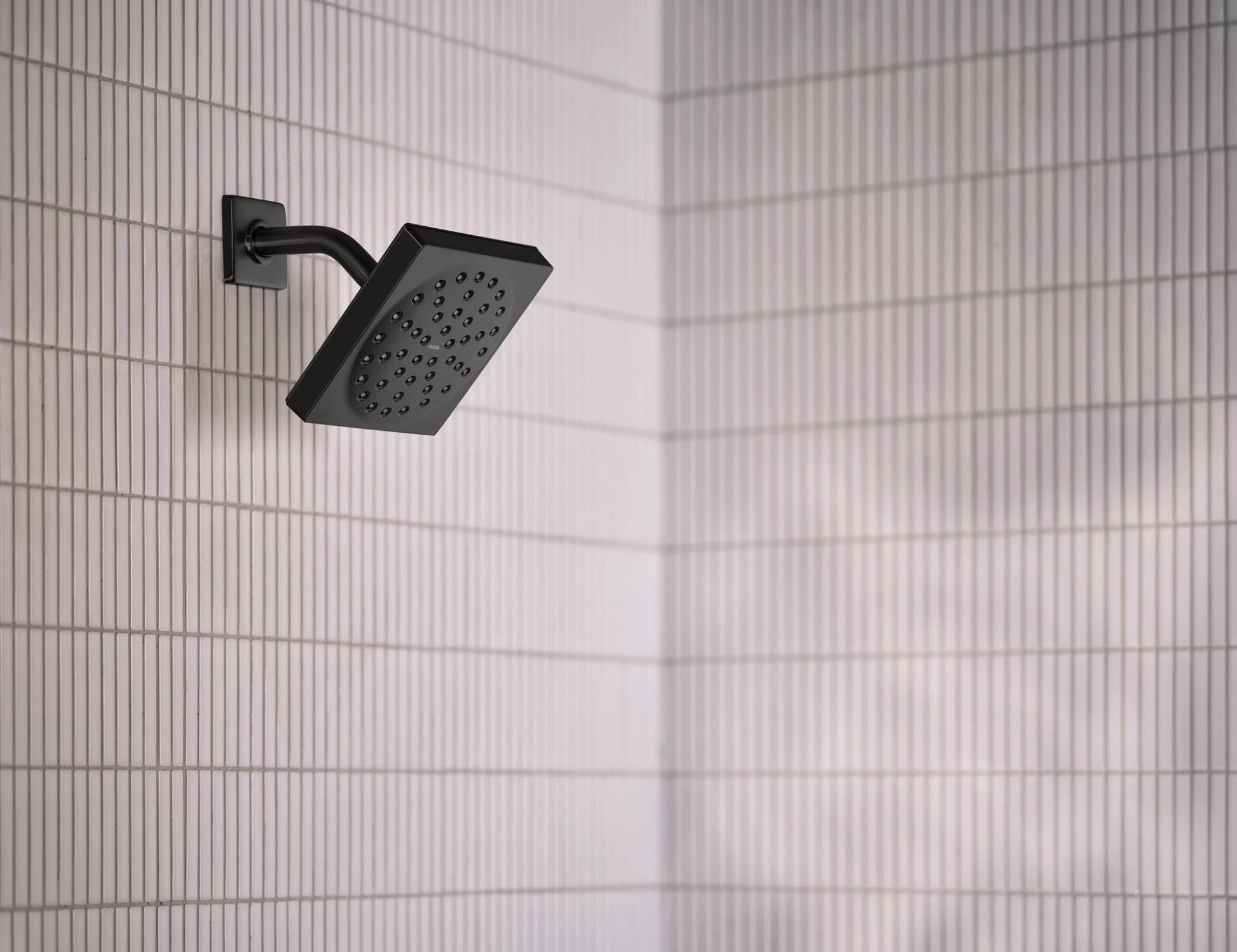 Matte black square shower head with perforated face mounted on curved arm against tiled wall.