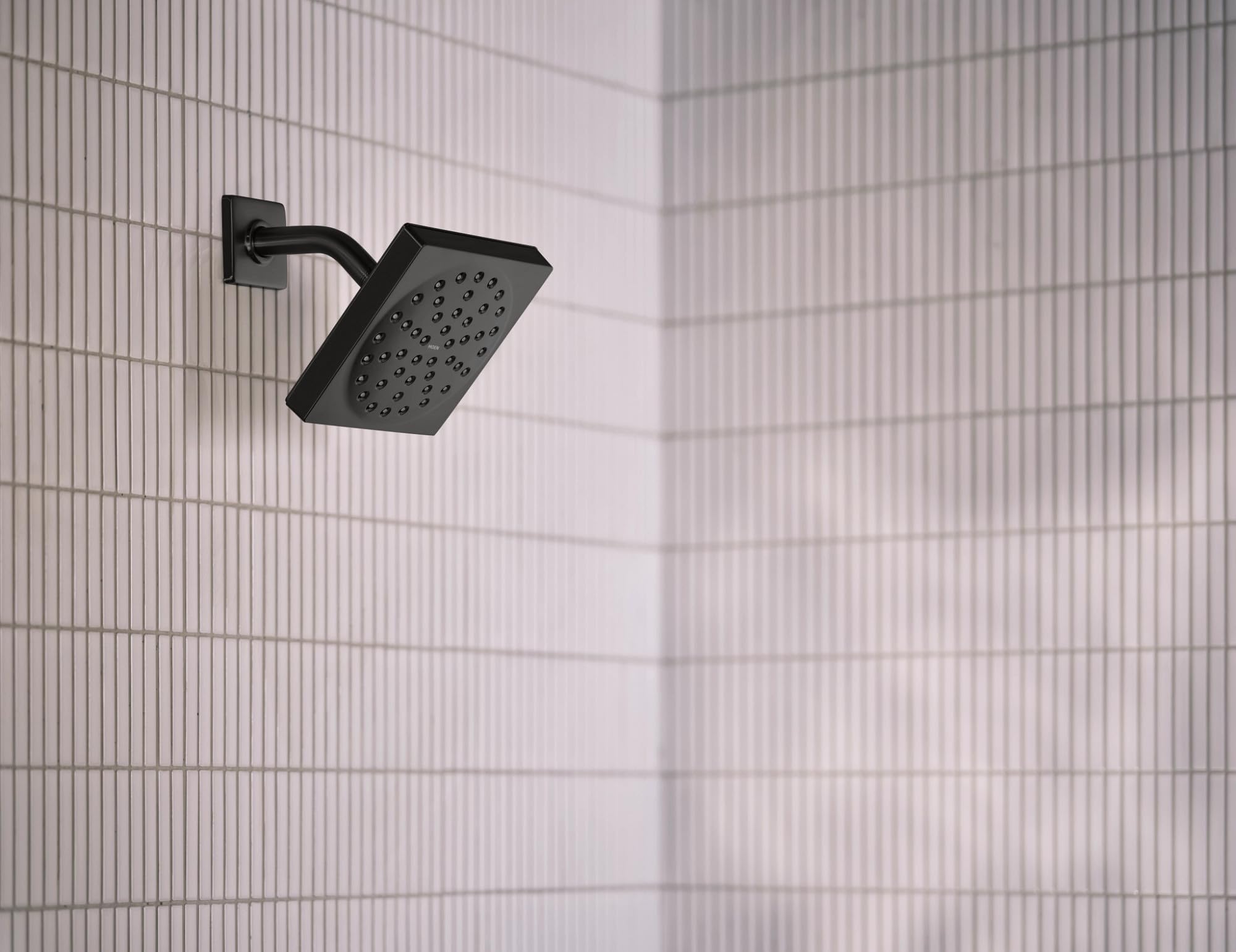 Matte black square shower head with perforated face mounted on curved arm against tiled wall.