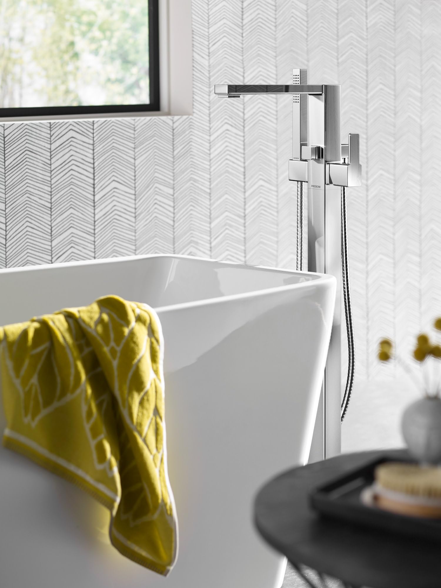 Brushed nickel floor-mount tub filler with single handle, angled spout, and attached handshower hose next to freestanding whi
