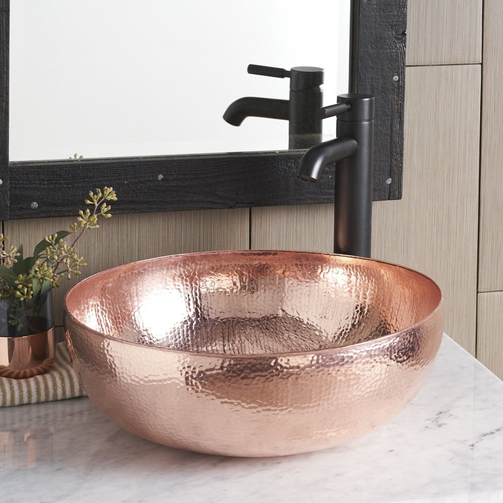 Polished copper round vessel sink with hammered texture and black faucet on marble countertop.
