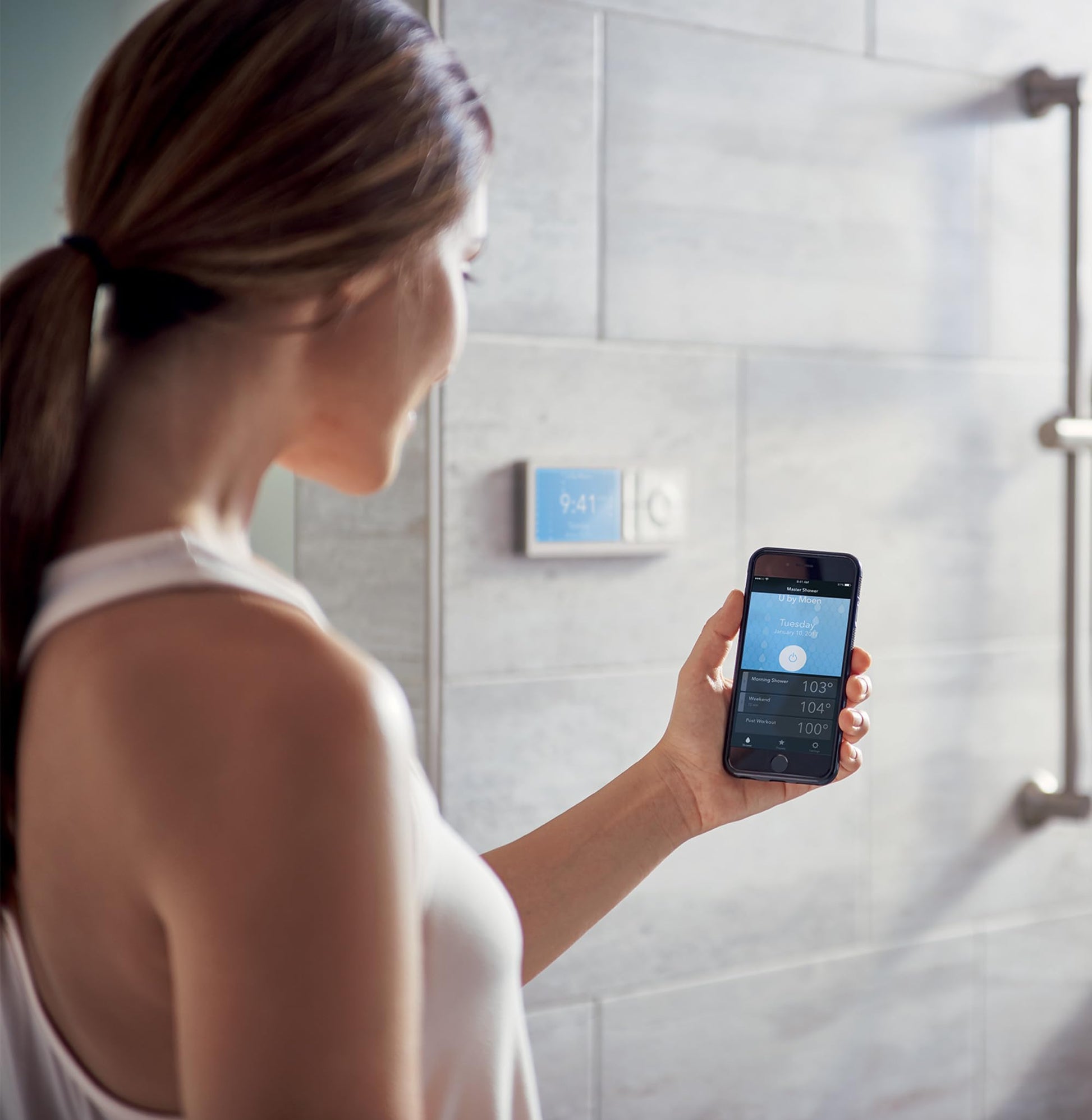 Person holding smartphone displaying shower control app with digital wall controller visible on tiled bathroom wall.