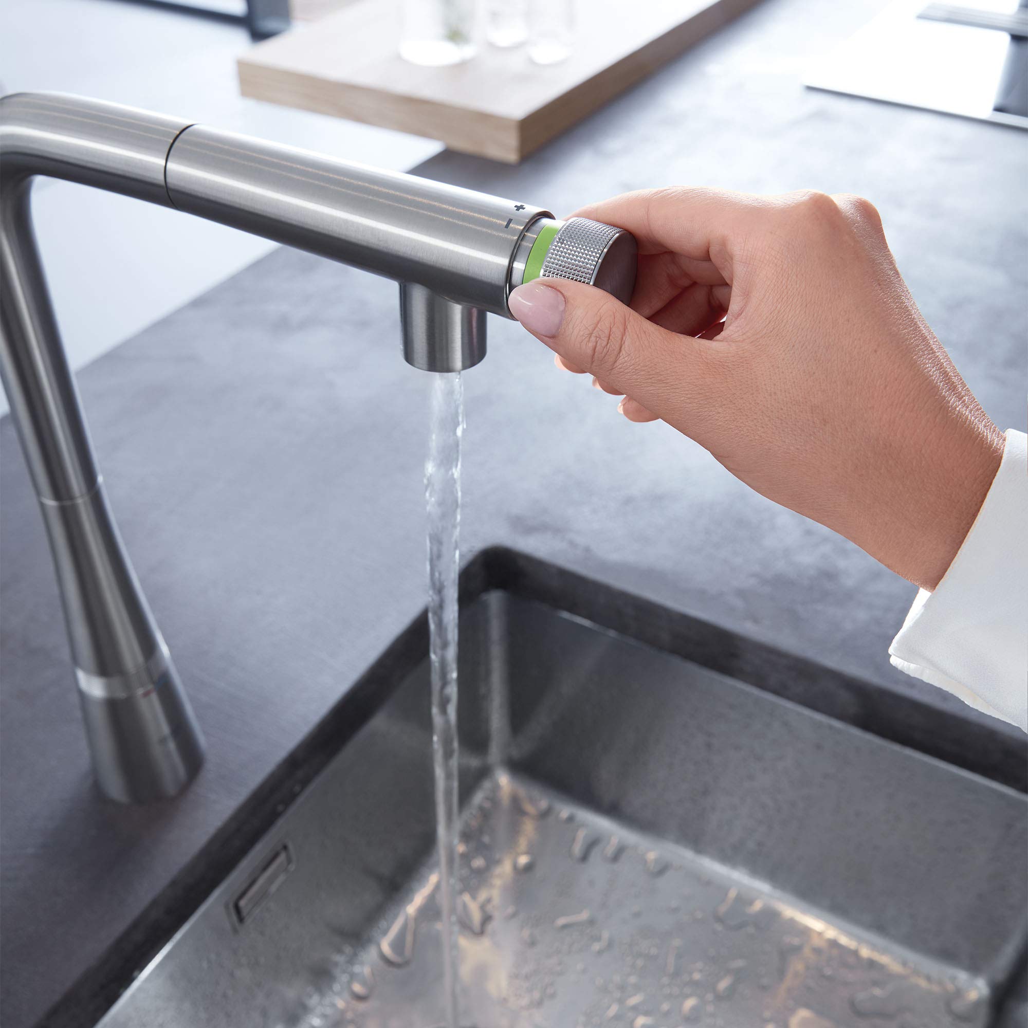 Stainless steel pull-out kitchen faucet with green control ring being operated over a sink with water flowing.