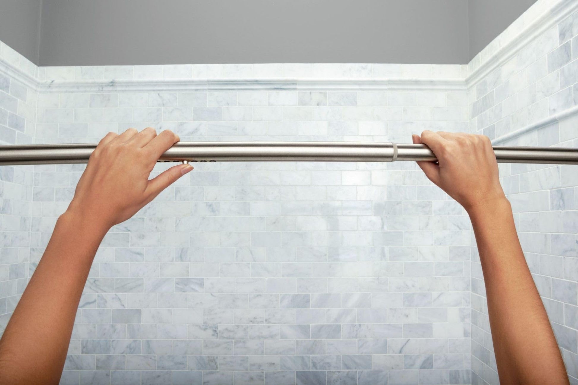 Brushed nickel curved tension shower curtain rod installed in bathroom with marble tile walls.