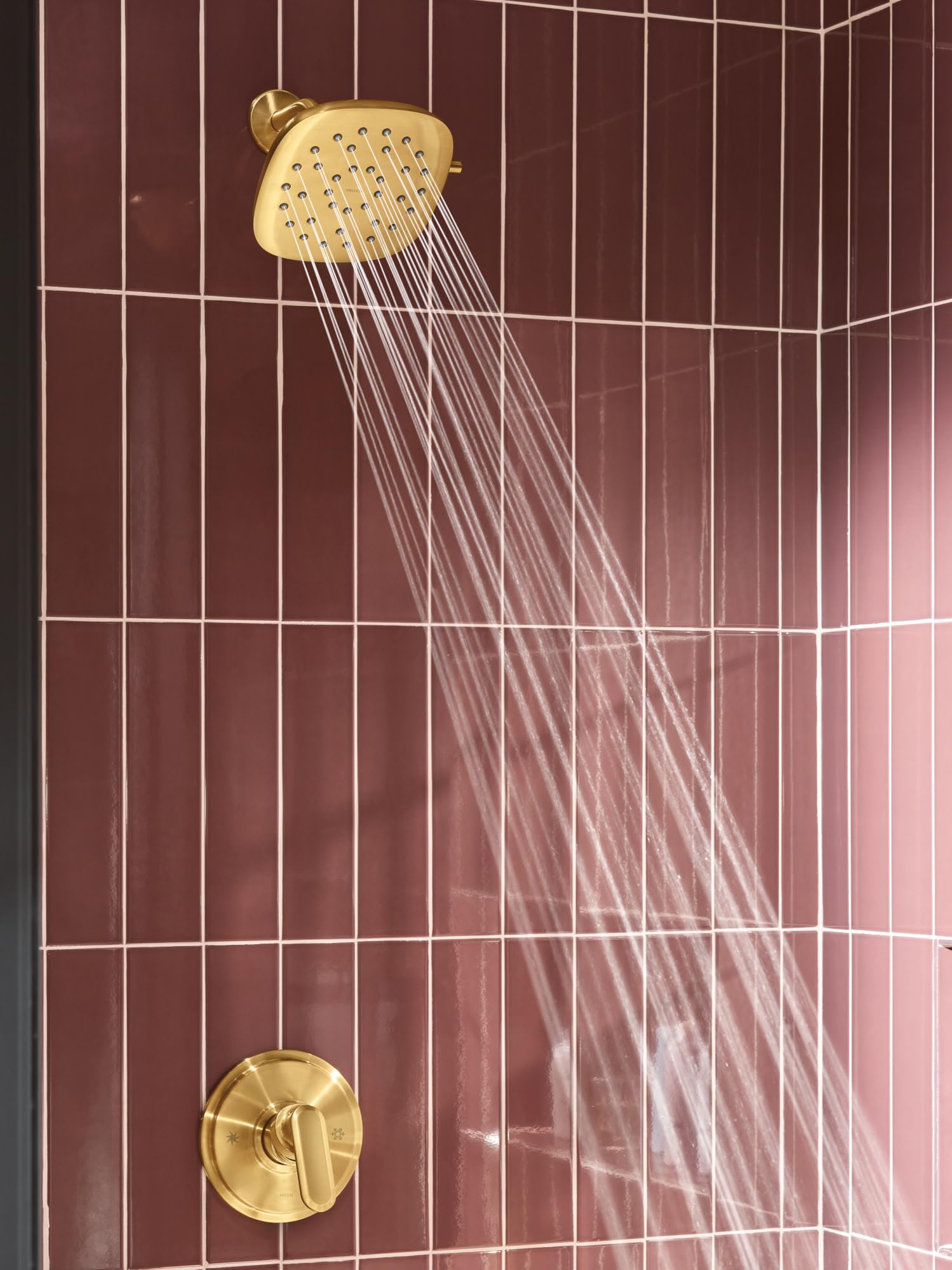 Brushed gold square rain shower head with multiple spray jets mounted on burgundy tiled bathroom wall.