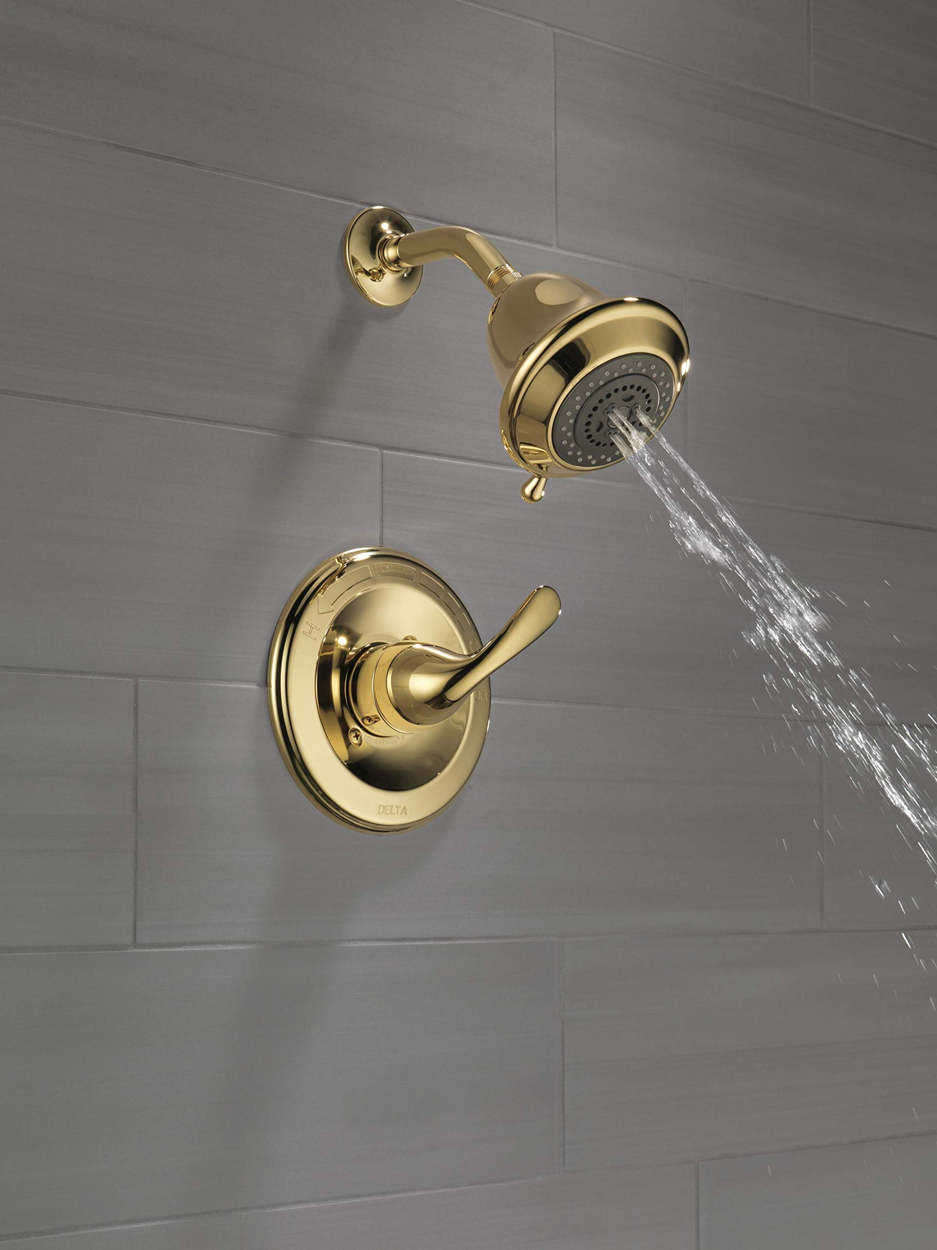 Polished brass shower trim with single lever handle and showerhead mounted on gray tile wall with water flowing.