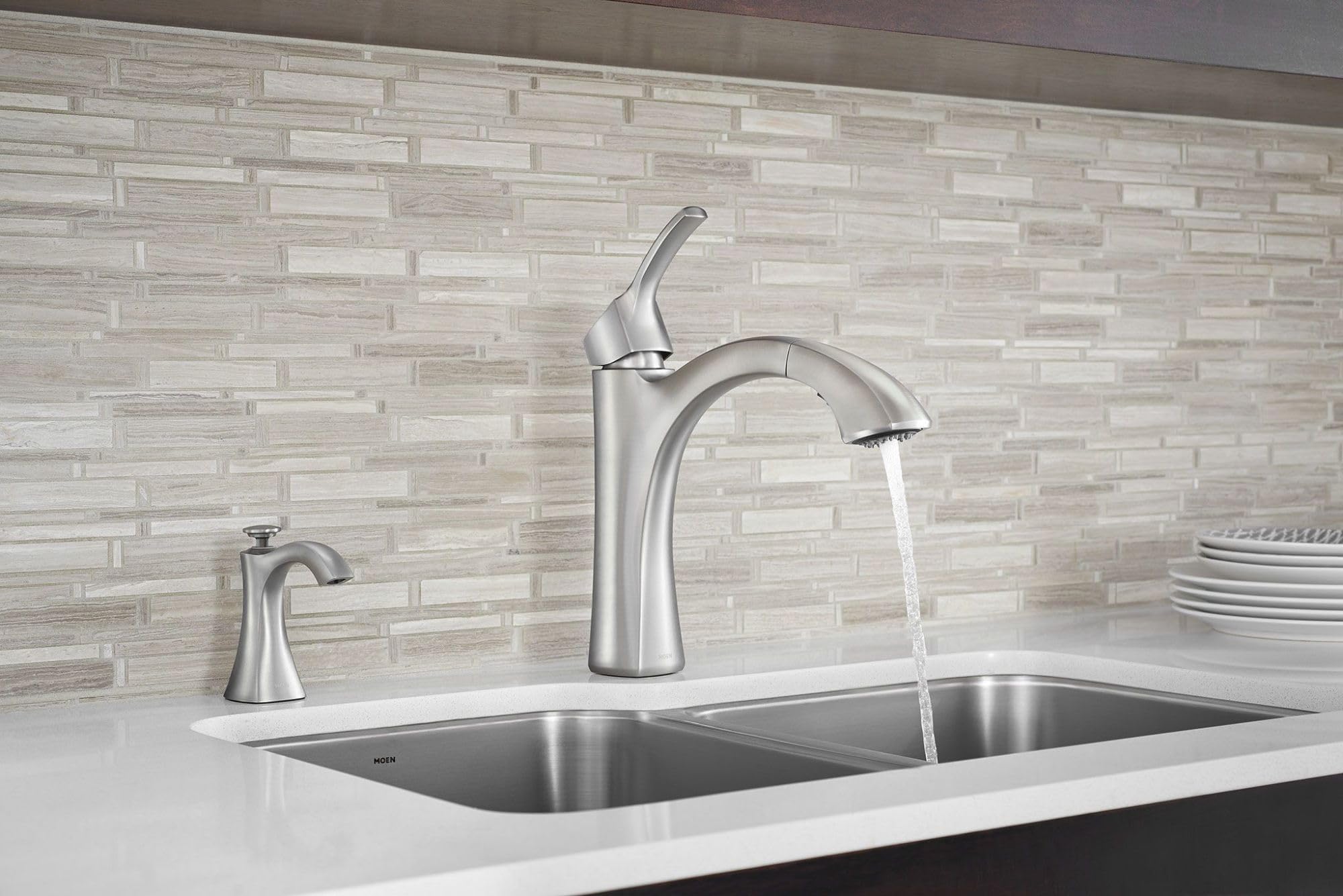 Stainless steel kitchen soap dispenser with curved spout mounted on white countertop beside faucet.