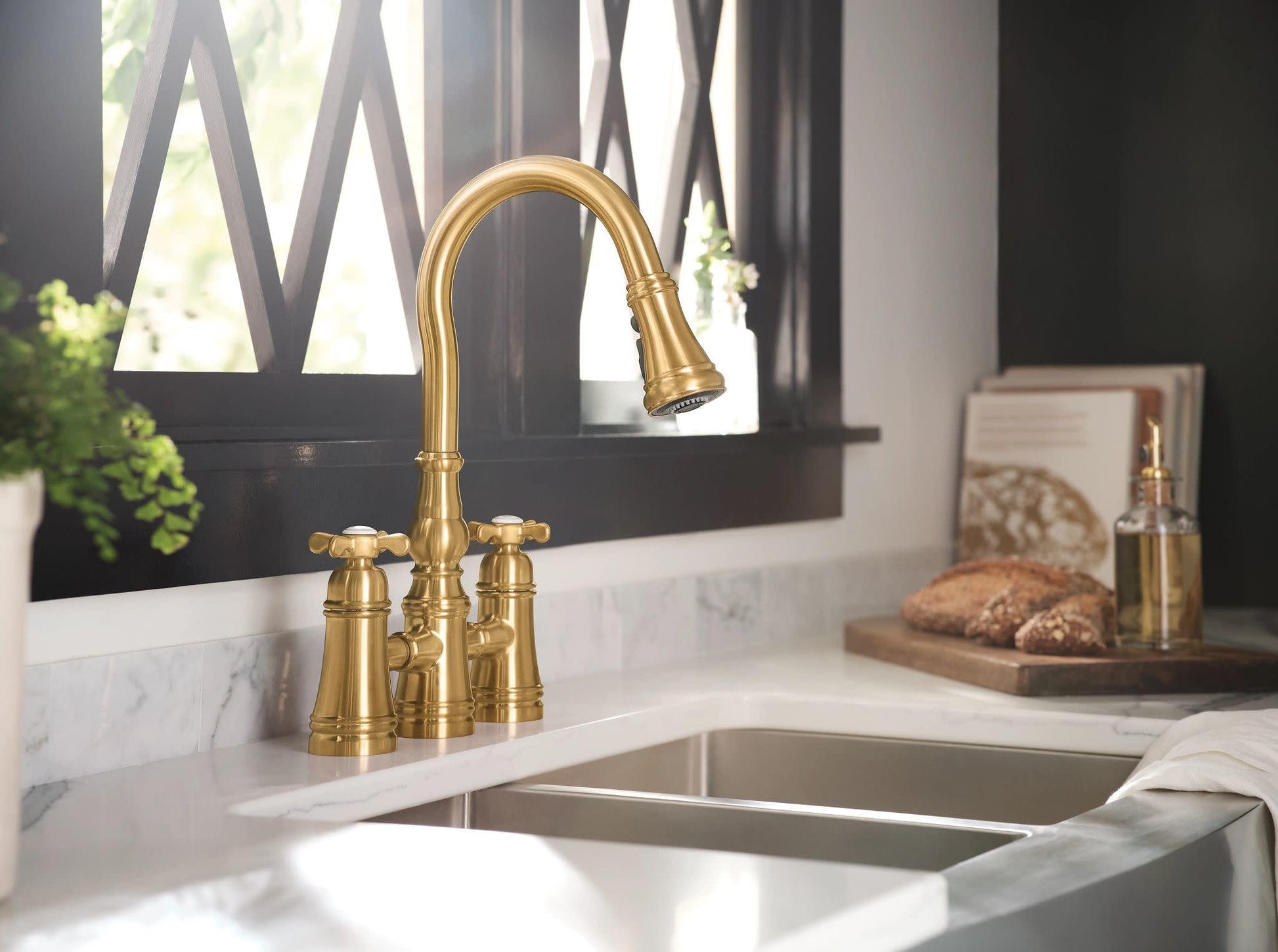 Brushed gold two-handle bridge kitchen faucet with pulldown spout over double basin sink.