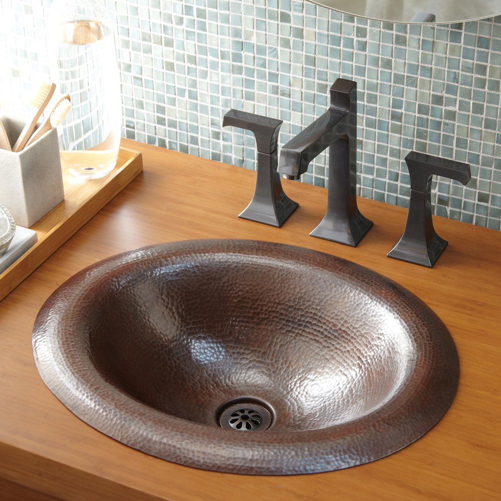 Antique copper oval bathroom sink with hammered finish and dark metal three-hole faucet on wooden vanity.