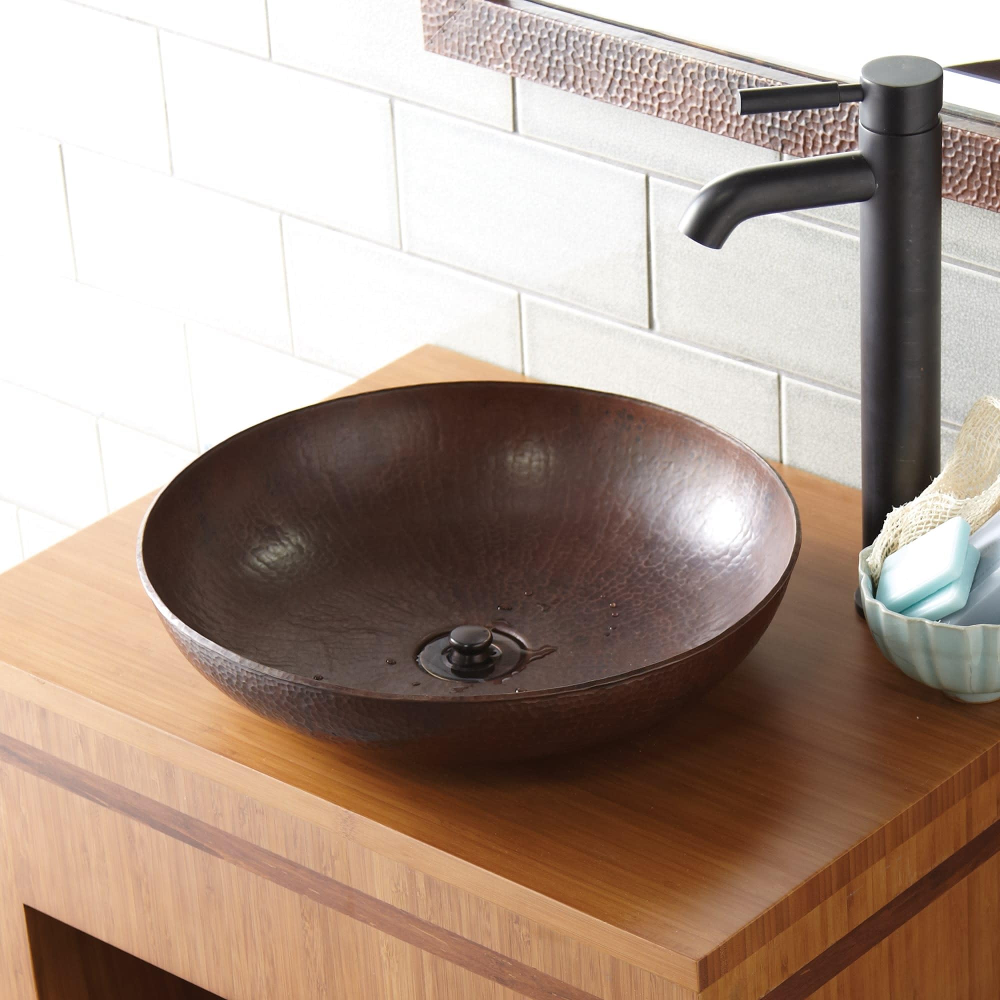 Hammered copper vessel sink with dark patina finish on wooden countertop, paired with black faucet.