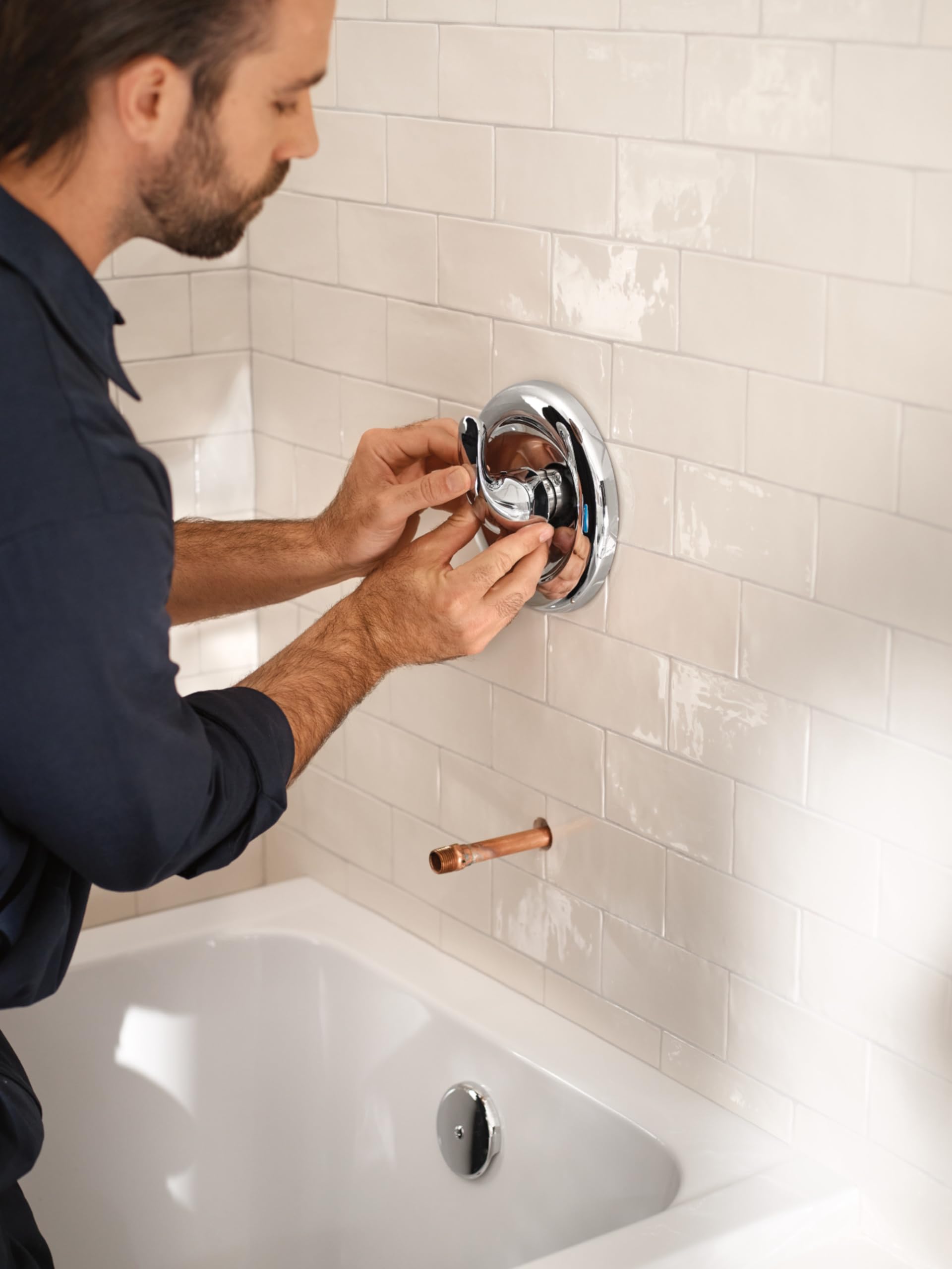 Chrome shower valve with trim ring being installed in white subway tile wall by a person.